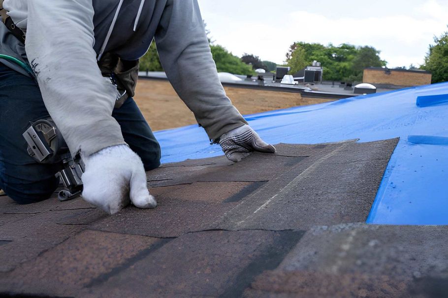 Worker installing shingles on a roof beside blue underlayment and gray roofing tools