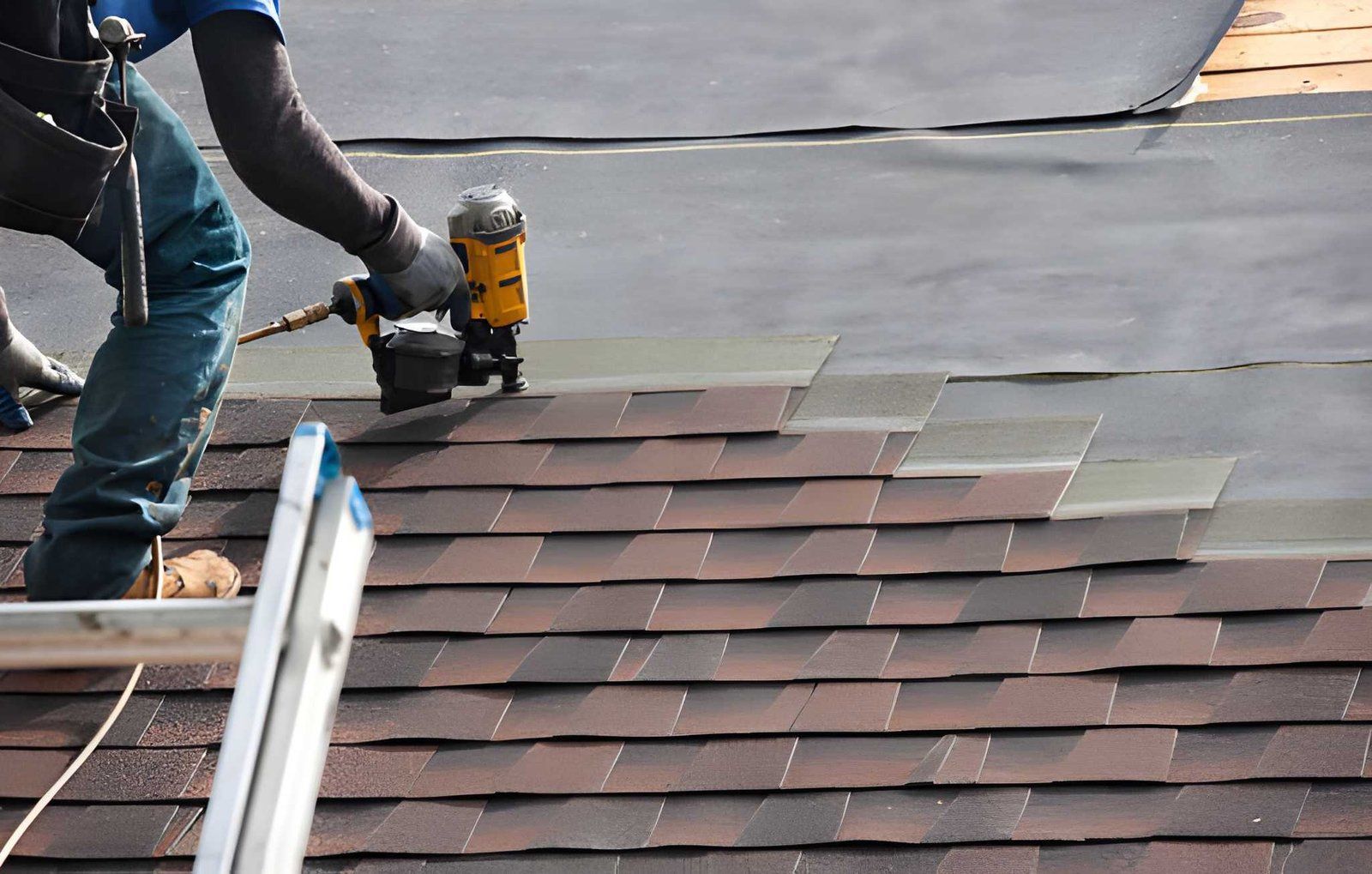 Worker using a nail gun to install shingles on a roof, with a ladder in the foreground