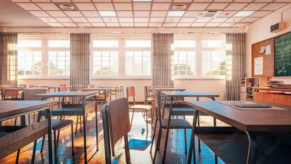 Empty sunlit classroom with desks, chairs, chalkboard, and windows.