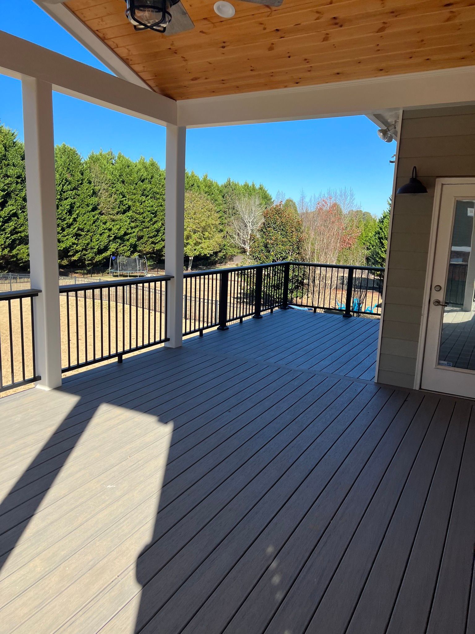 Covered deck with gray flooring, black railing, and a view of trees under a blue sky.