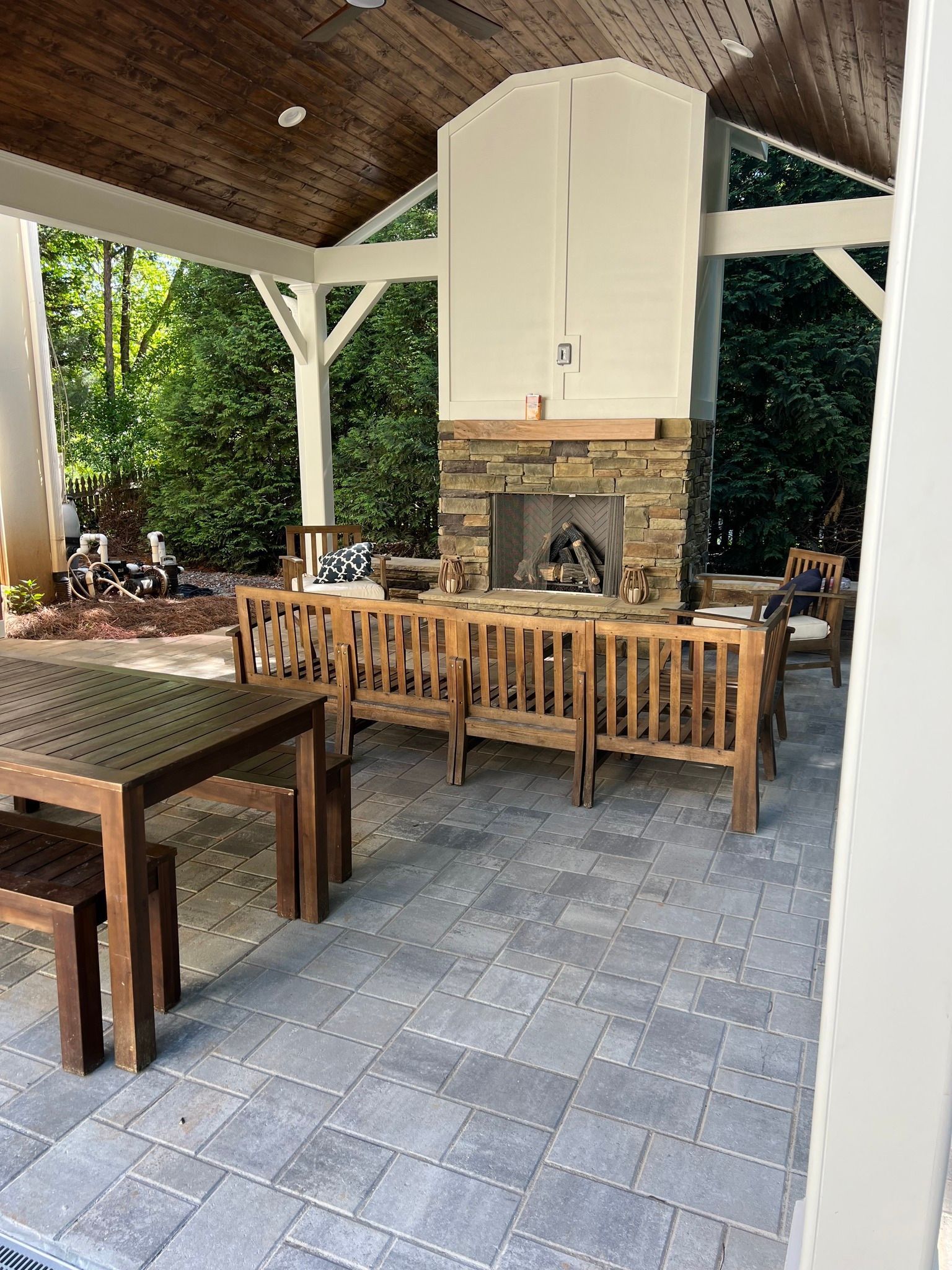 Covered patio with fireplace, stone patio, wooden furniture, and lush greenery.