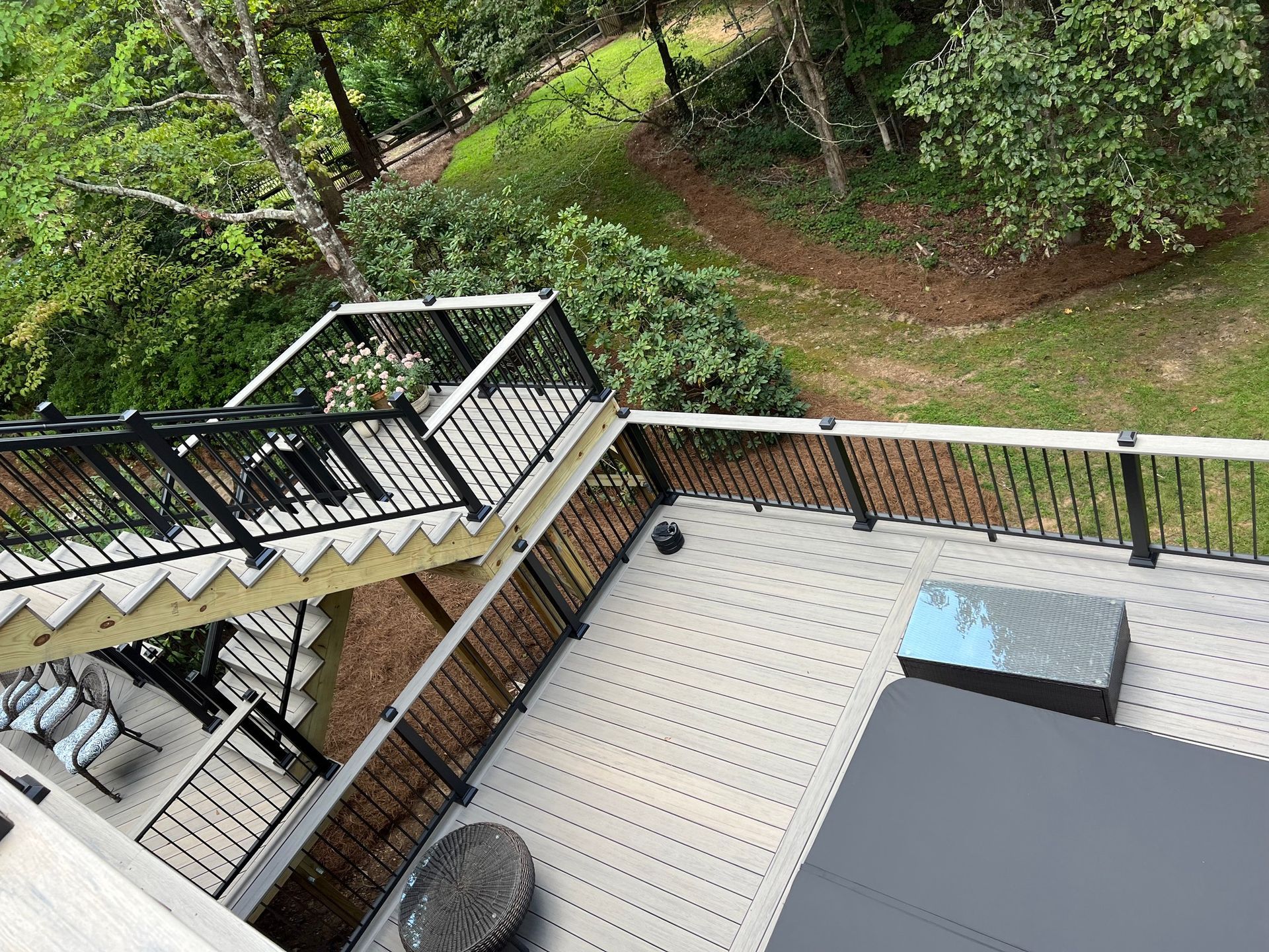 Deck with stairs, black railings, and gray composite decking. Lush green trees and foliage in background.