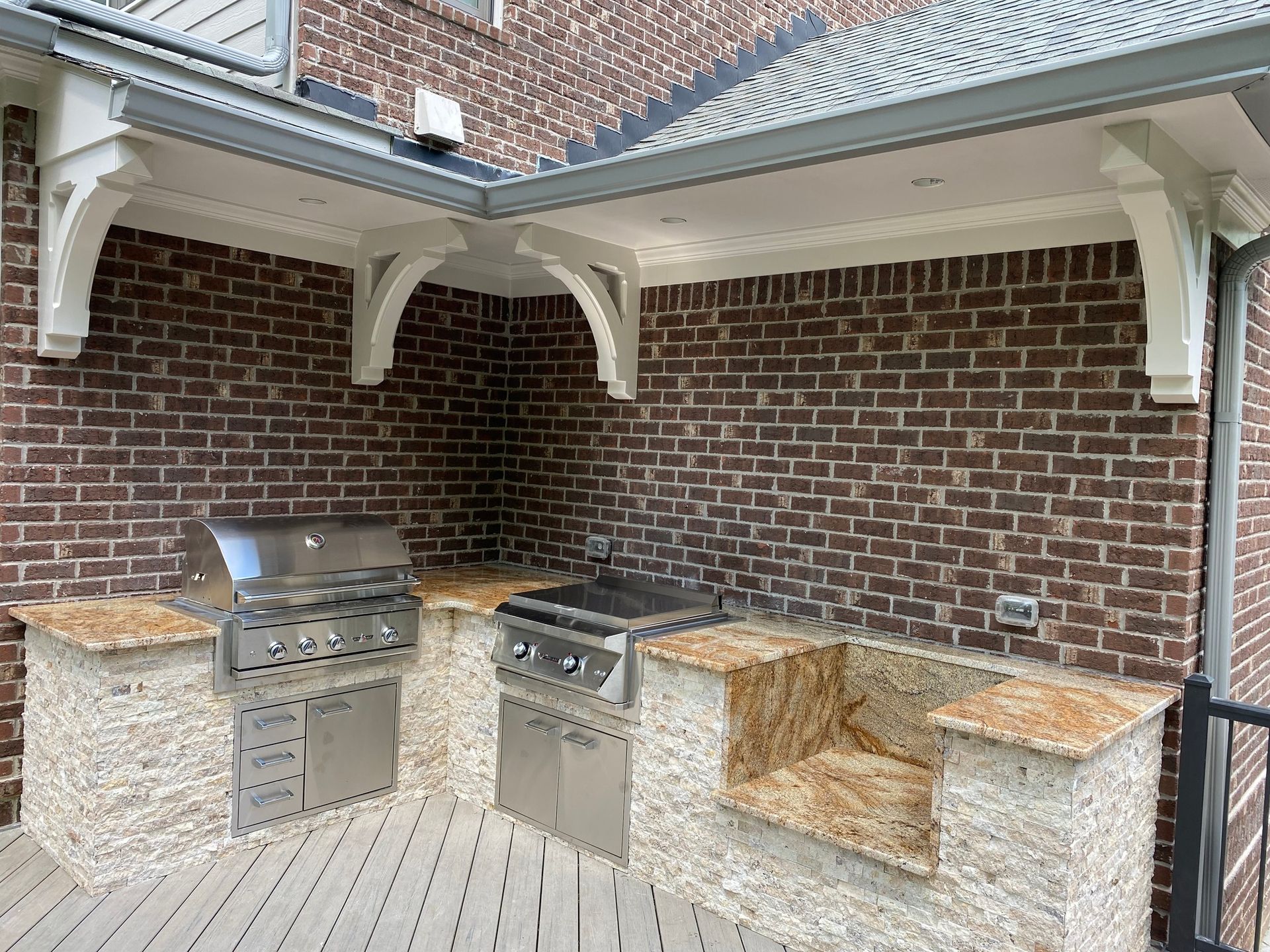 Outdoor kitchen with brick wall, granite countertops, and stainless steel grills under a covered roof.