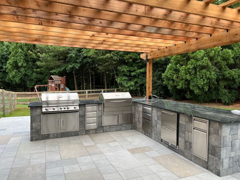 Outdoor kitchen with grill, sink, and refrigerator under a wooden pergola on a stone patio.