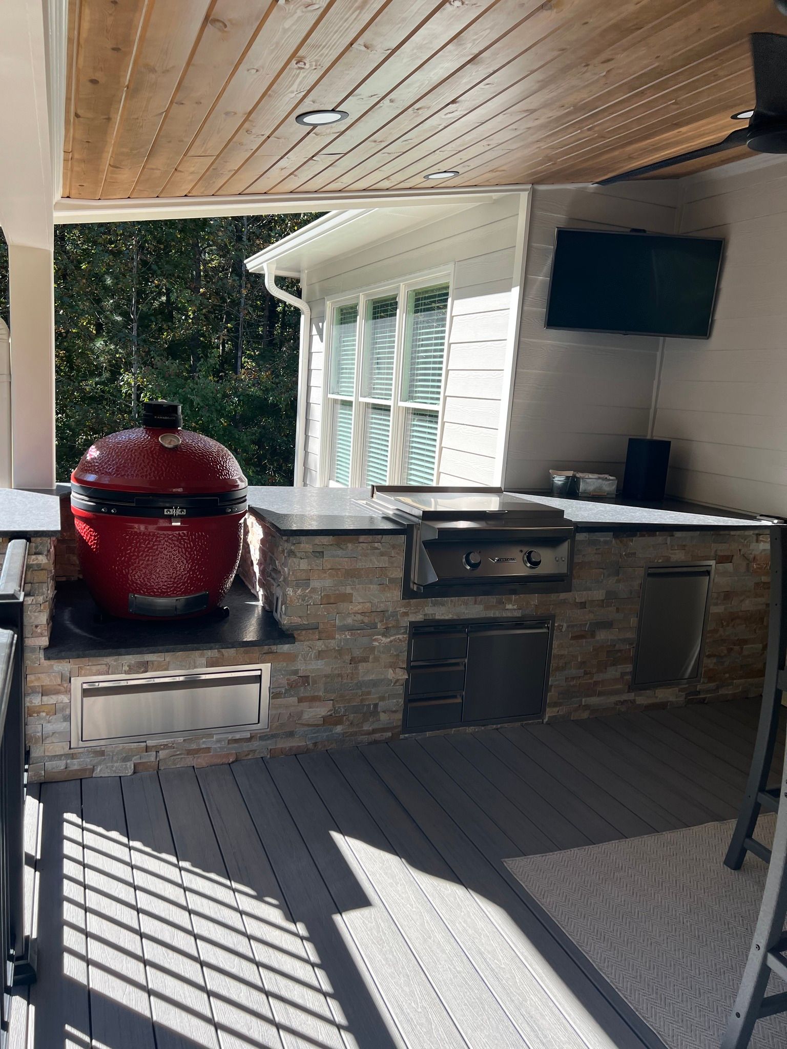 Outdoor kitchen with red grill, built-in appliances, stone facade, and TV on a patio.