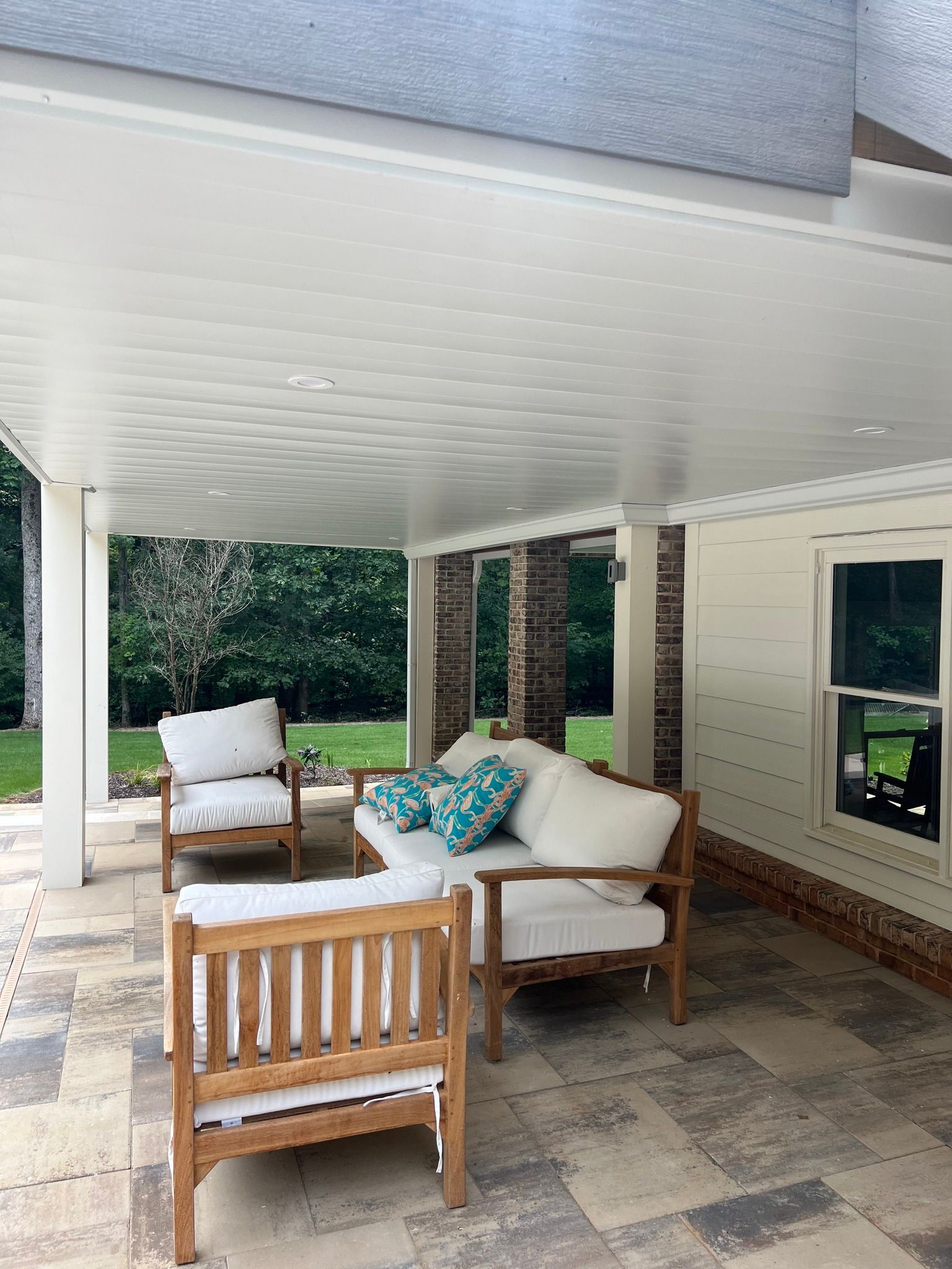 Patio seating under a white roof. Teak furniture with white cushions. Brick and cream-colored wall.