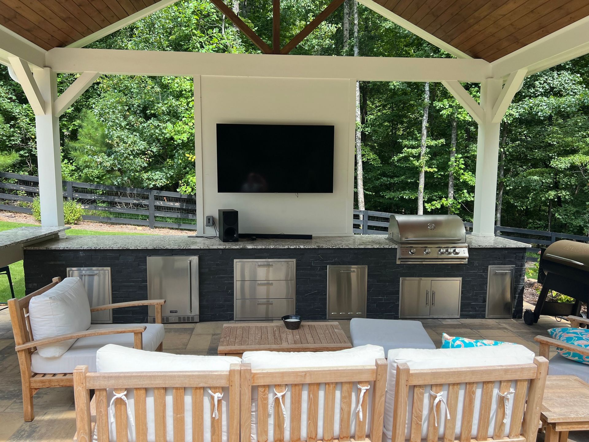 Outdoor kitchen under a wooden canopy with a grill, cabinets, and a mounted TV.
