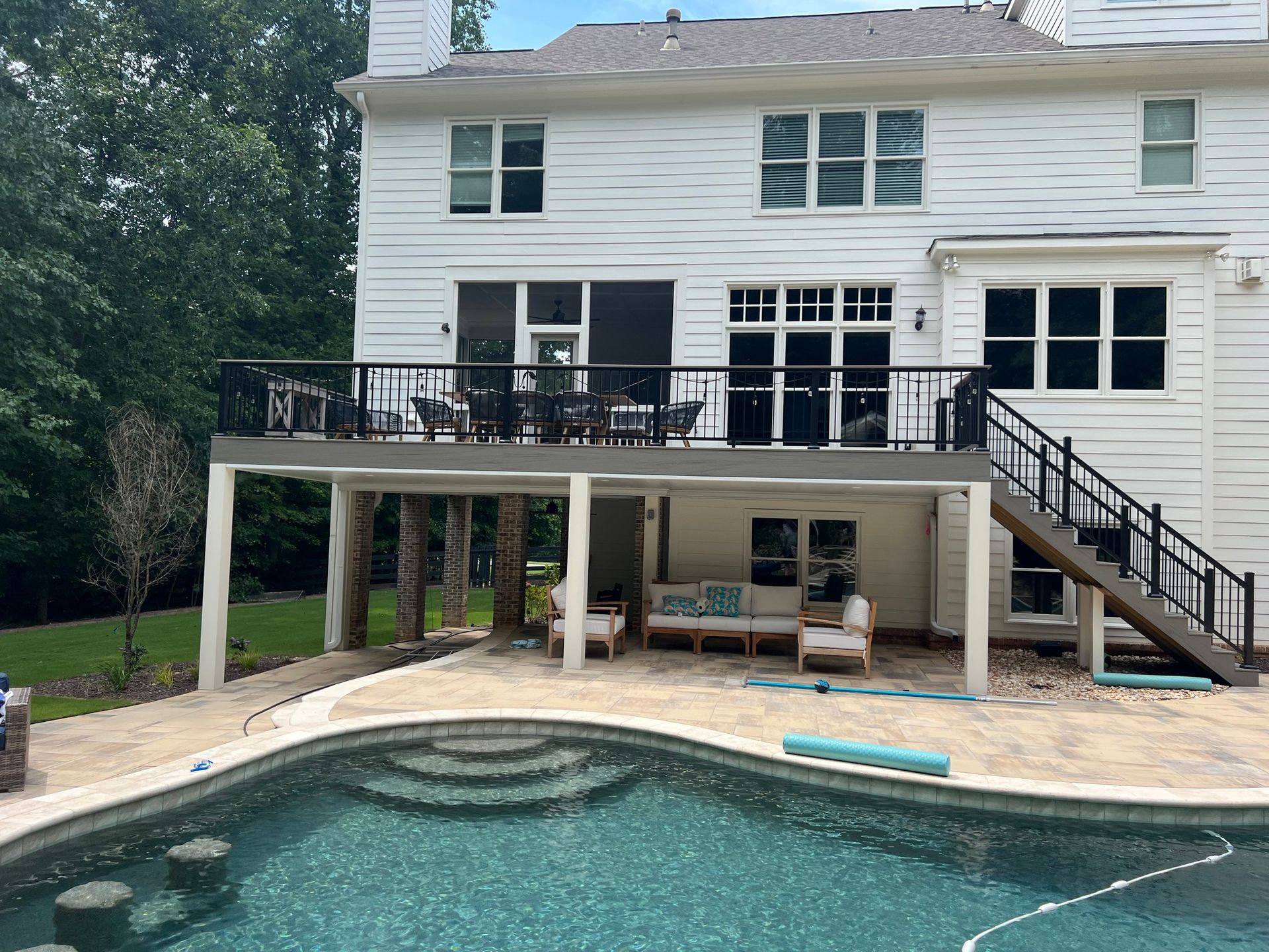 Two-story white house with a deck overlooking a pool. Outdoor living area with furniture beneath the deck.