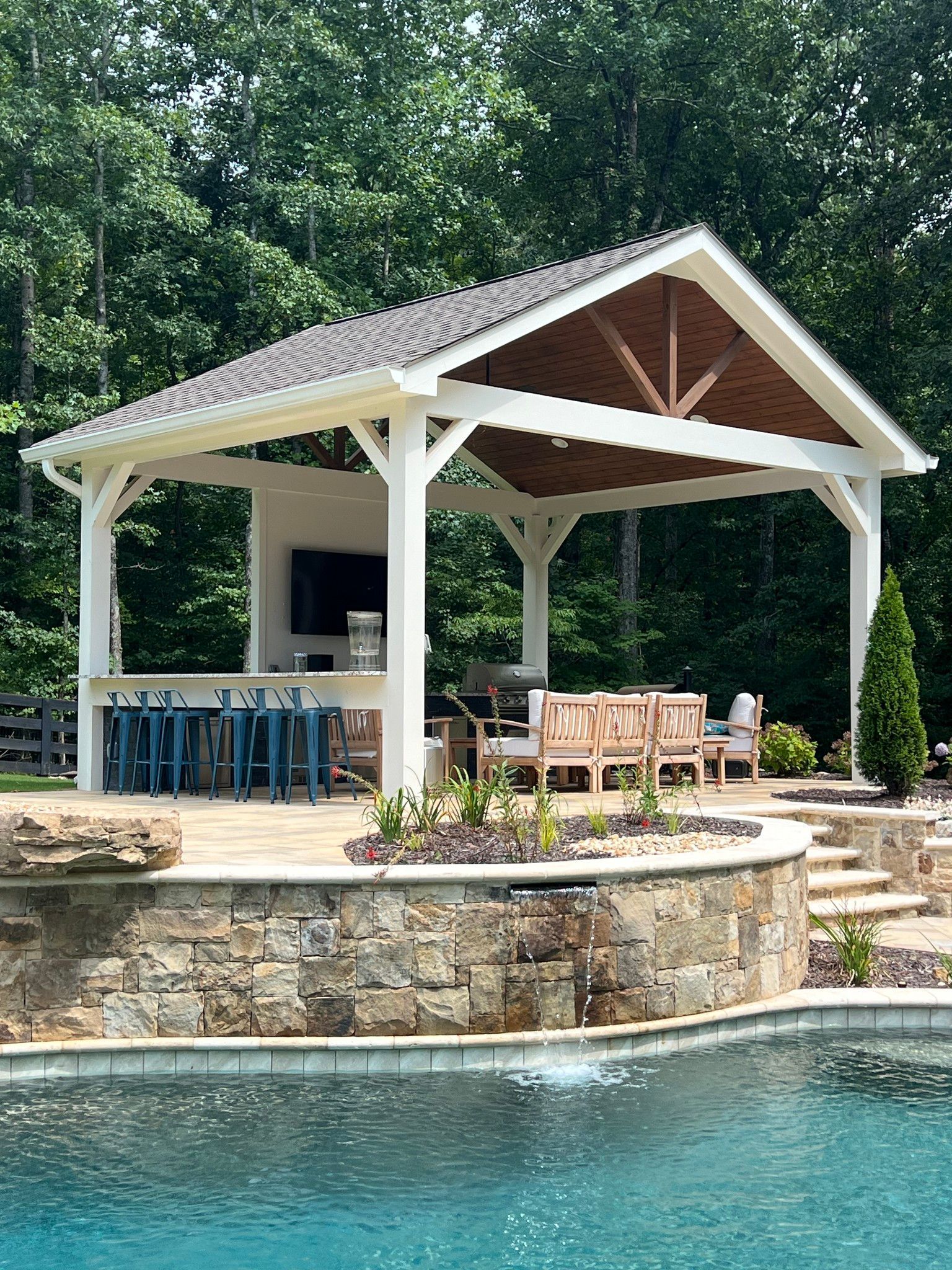 Poolside pavilion with bar, seating, and grill. White wood frame, blue stools, stone wall, and trees in background.