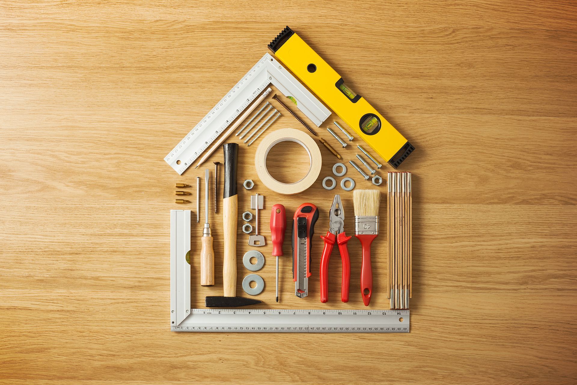 Tools arranged in the shape of a house on a wooden surface, including a level, tape, and pliers.