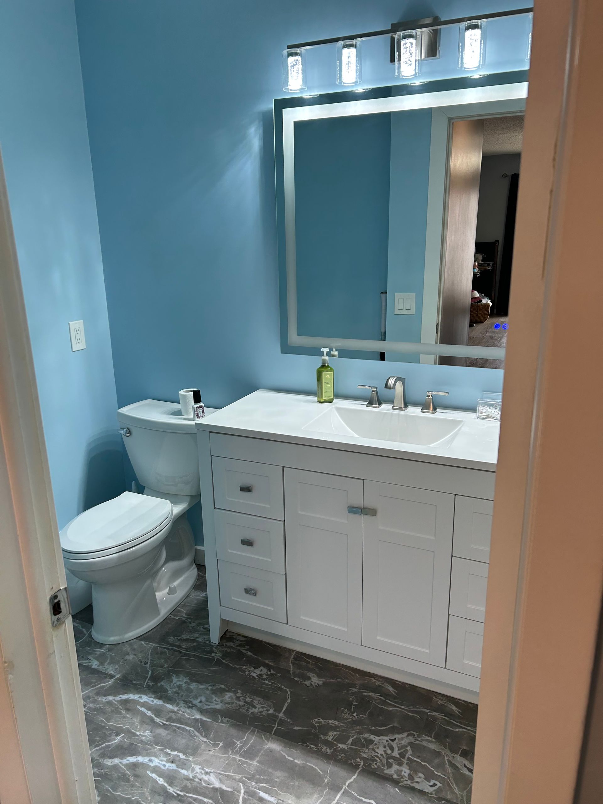 Bathroom with white vanity, blue walls, and gray tile floor. Mirror with lights above the double sinks.
