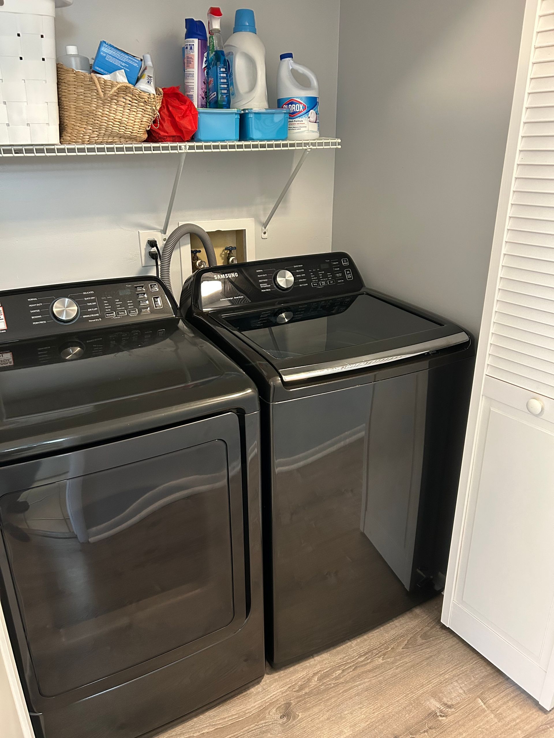 Black washer and dryer in a laundry room with cleaning supplies on a shelf.