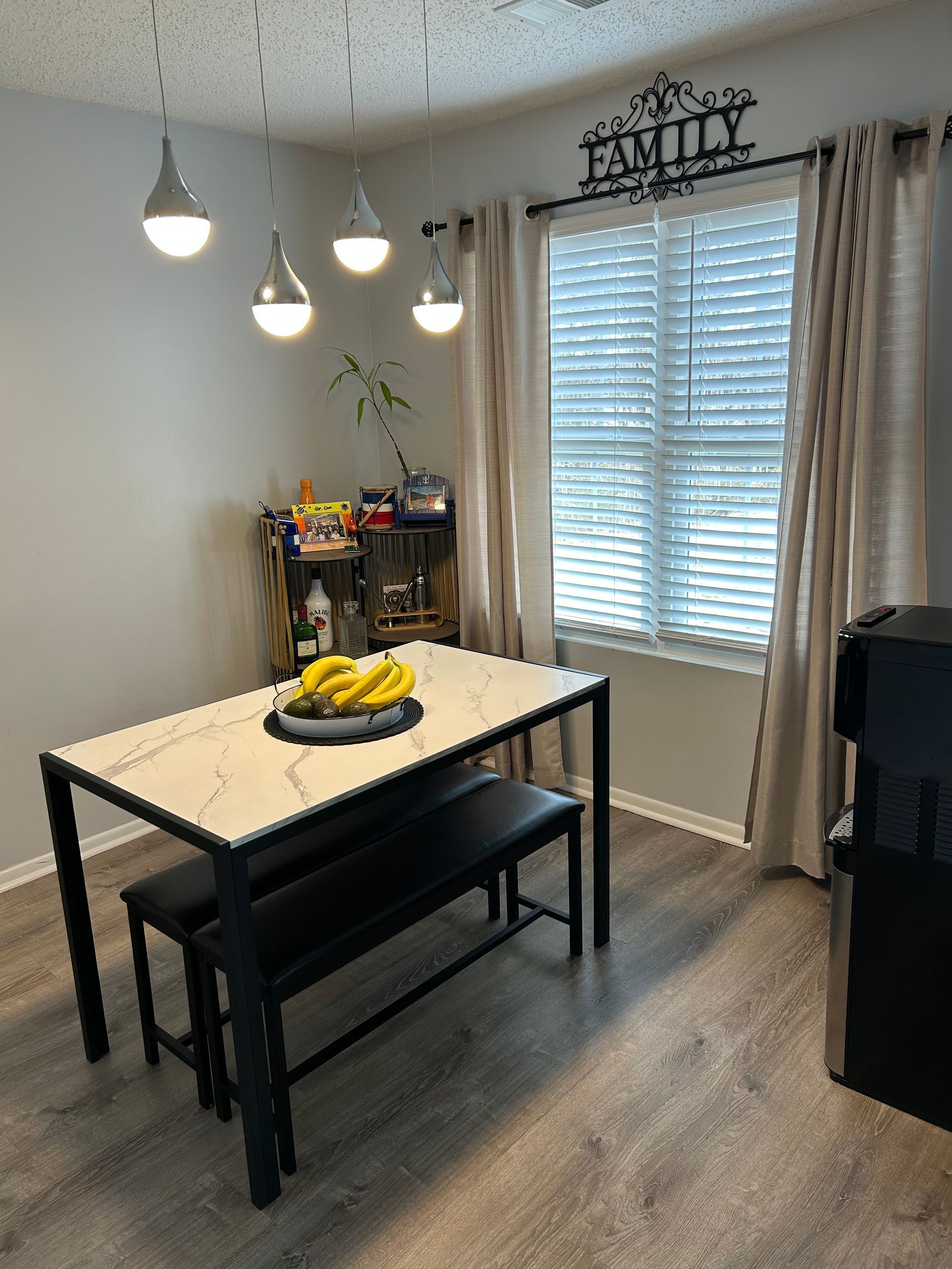 Dining room with a table, benches, and a serving cart. Window with blinds and a family sign.