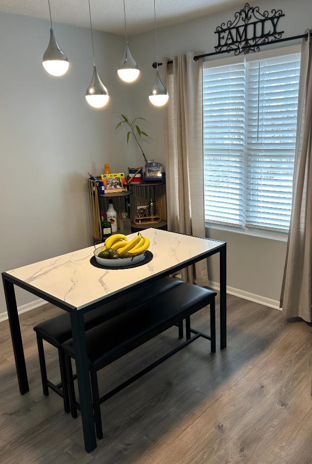 Dining room with table and benches, pendant lights, bar cart, window with blinds and beige curtains.