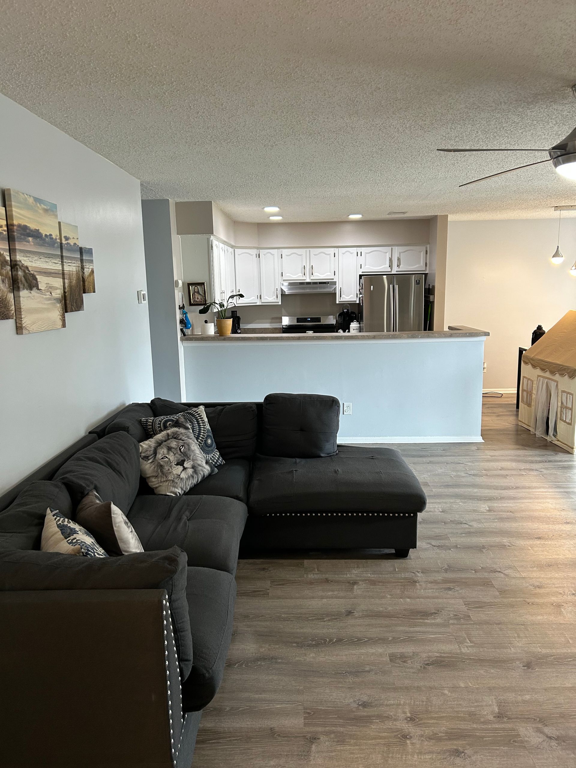 Living room with gray sectional sofa, open to a white-walled kitchen. Light wood flooring.