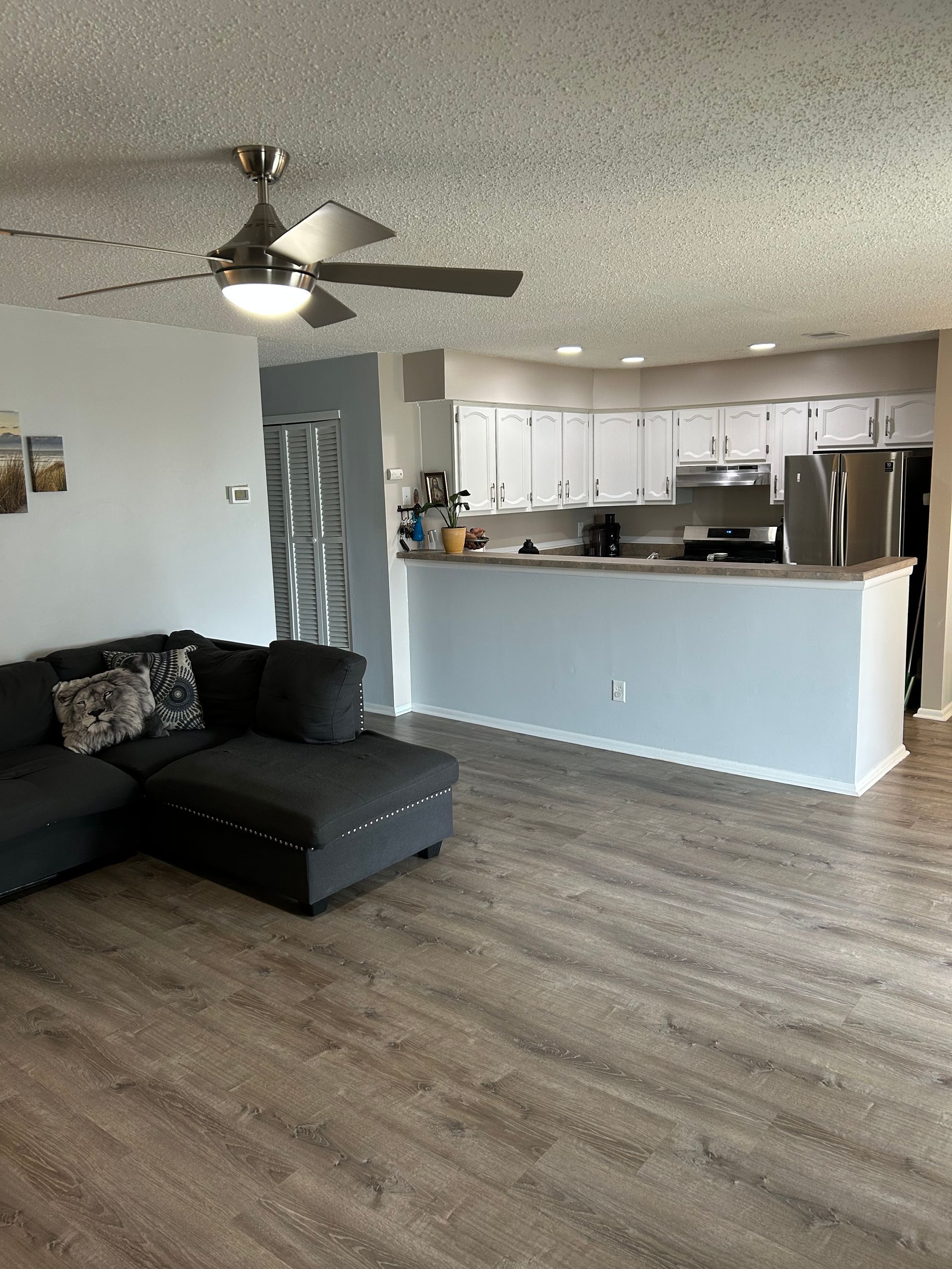 Living room with gray sectional, open to a white-cabinet kitchen with light gray walls and wood-look flooring.