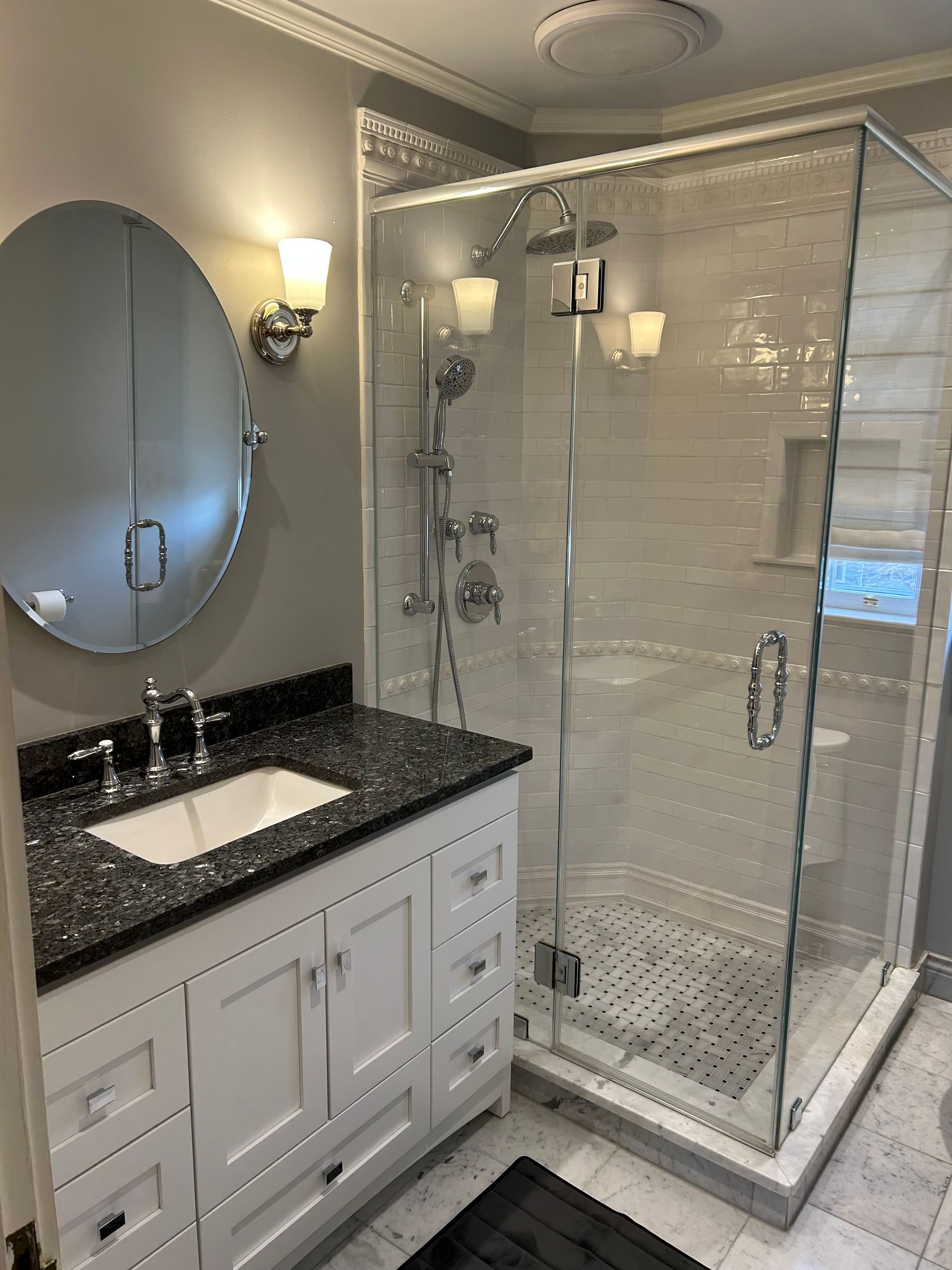 Bathroom with white vanity, black granite countertop, glass shower, and oval mirror.
