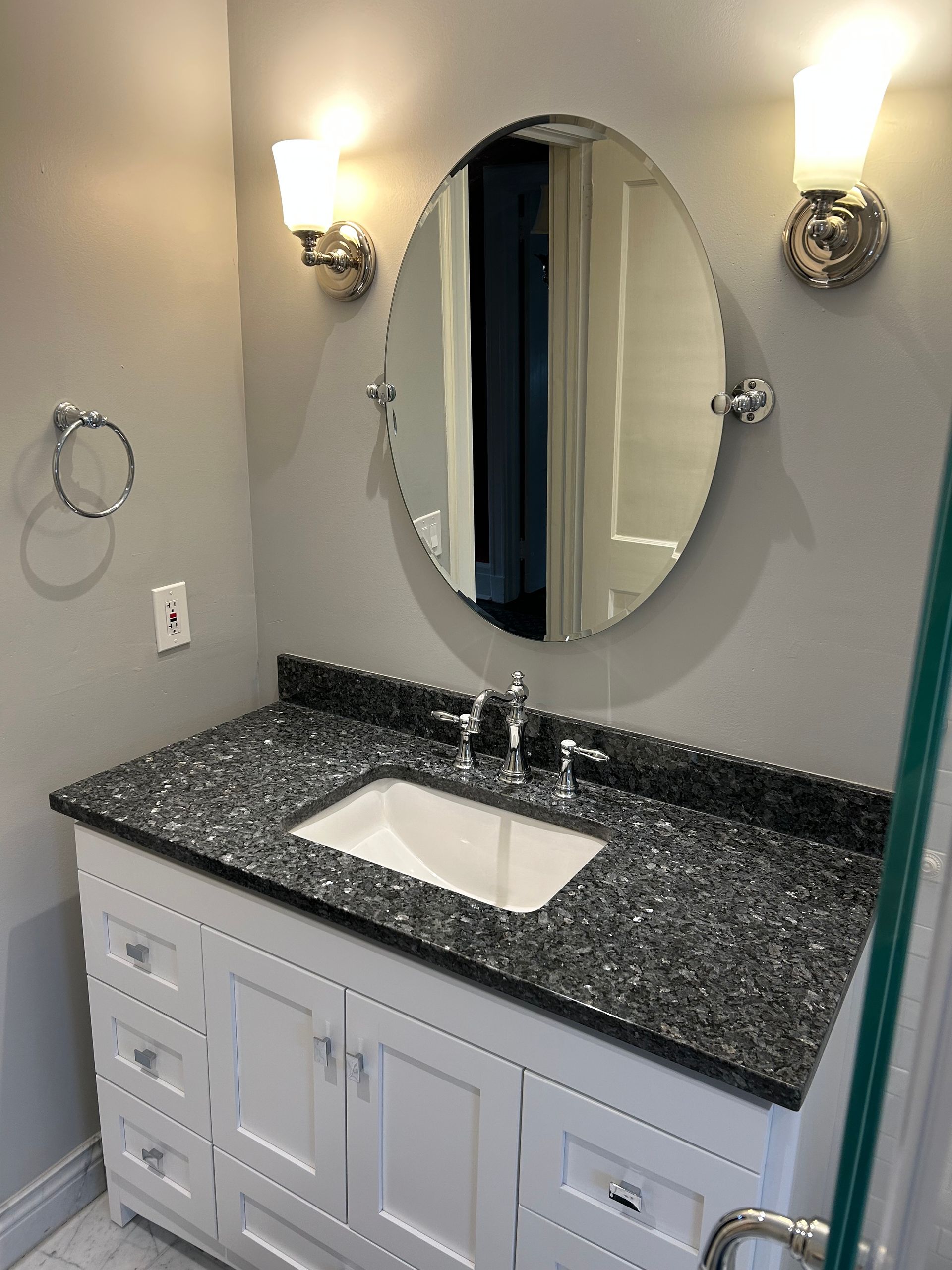 Bathroom vanity with oval mirror, granite countertop, white cabinets, and sconce lighting.