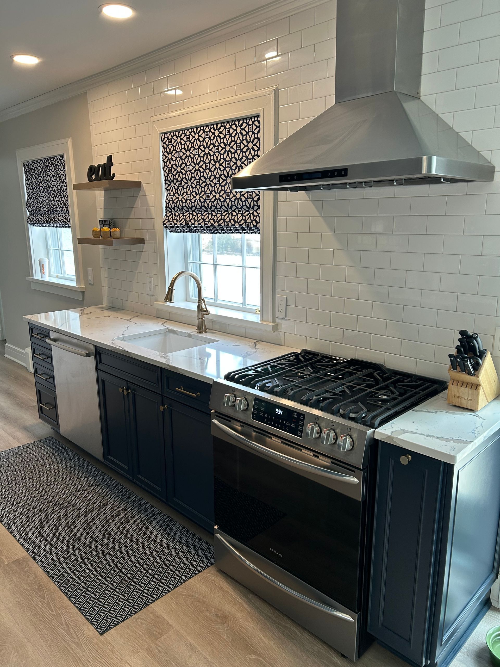 Modern kitchen with white countertops, navy cabinets, stainless steel appliances, and patterned window shades.