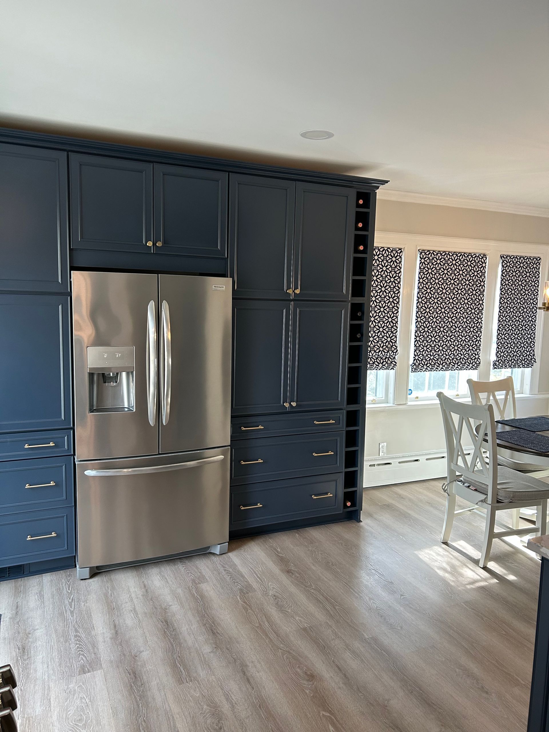 A kitchen with blue cabinets and a stainless steel refrigerator. A dining table with white chairs and a patterned window.