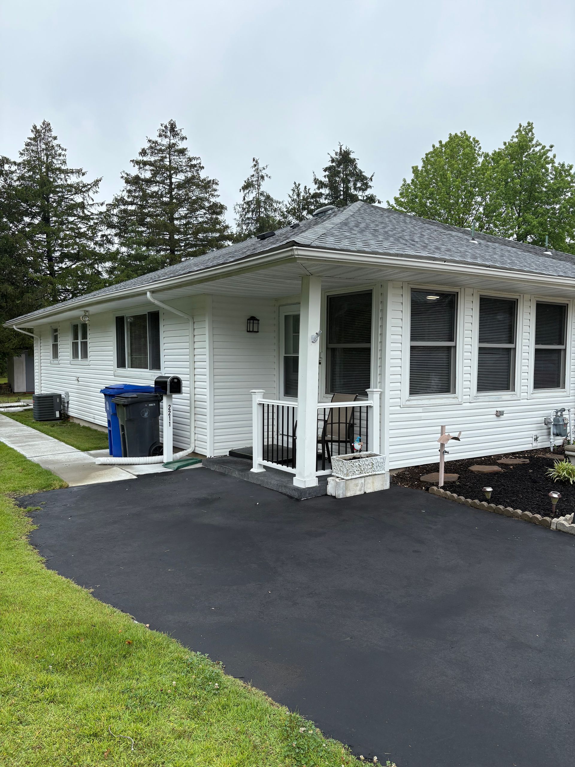 White house with black asphalt driveway and green grass lawn under a cloudy sky.
