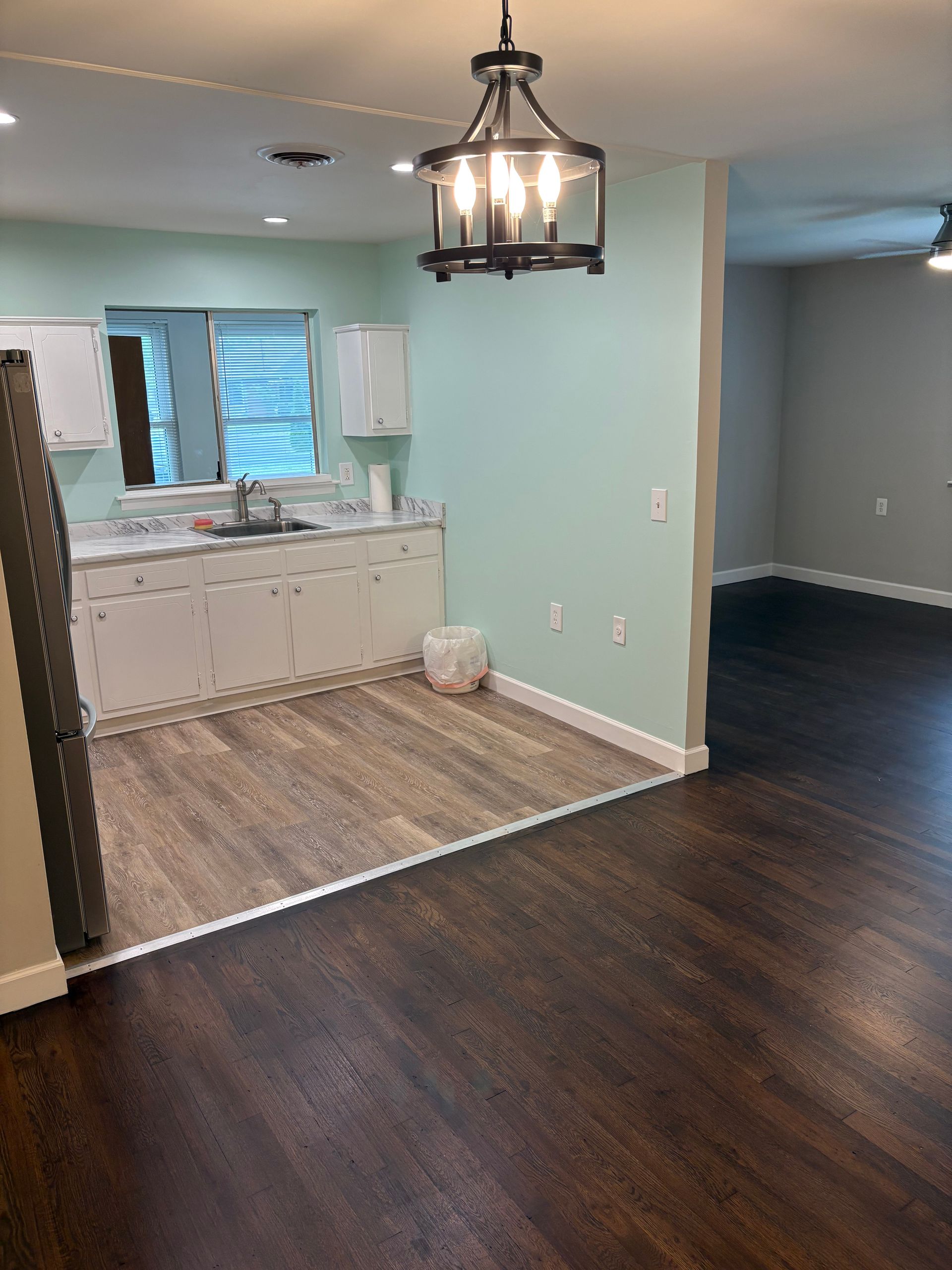 Interior with kitchen and living space, showing wood floors, light blue walls, and a hanging light fixture.
