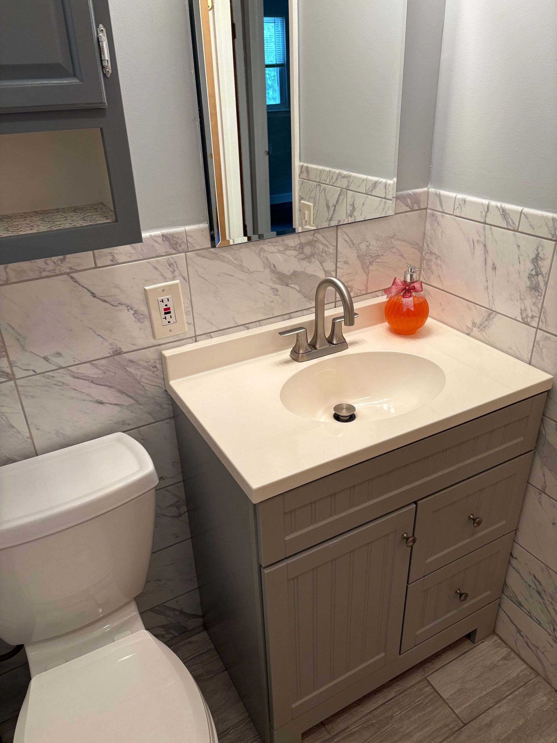 Bathroom with gray vanity, sink, toilet, and marble-look tiled walls. A mirror and overhead cabinet are visible.