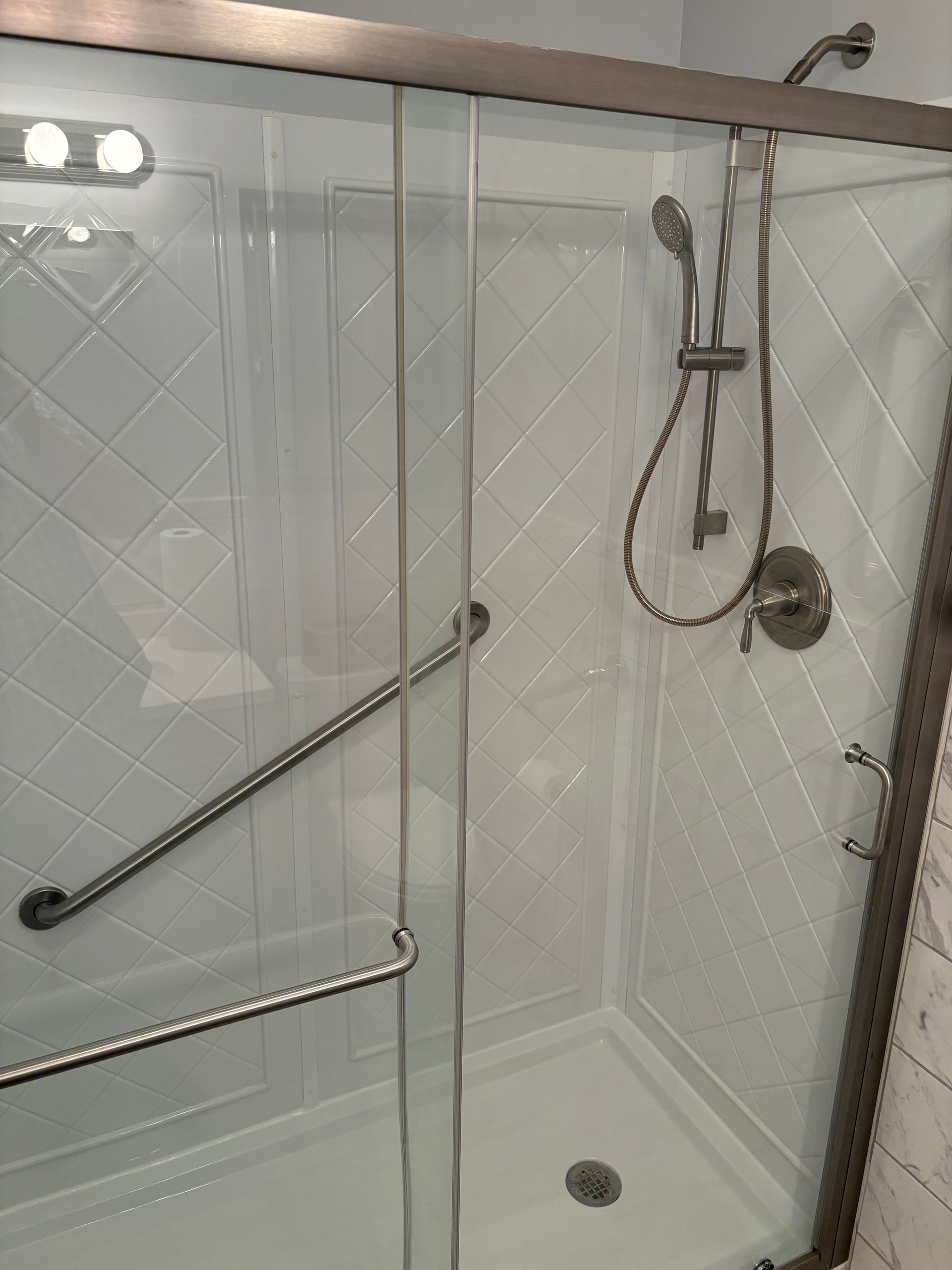 Accessible shower with grab bars and handheld showerhead. White tile and glass doors.