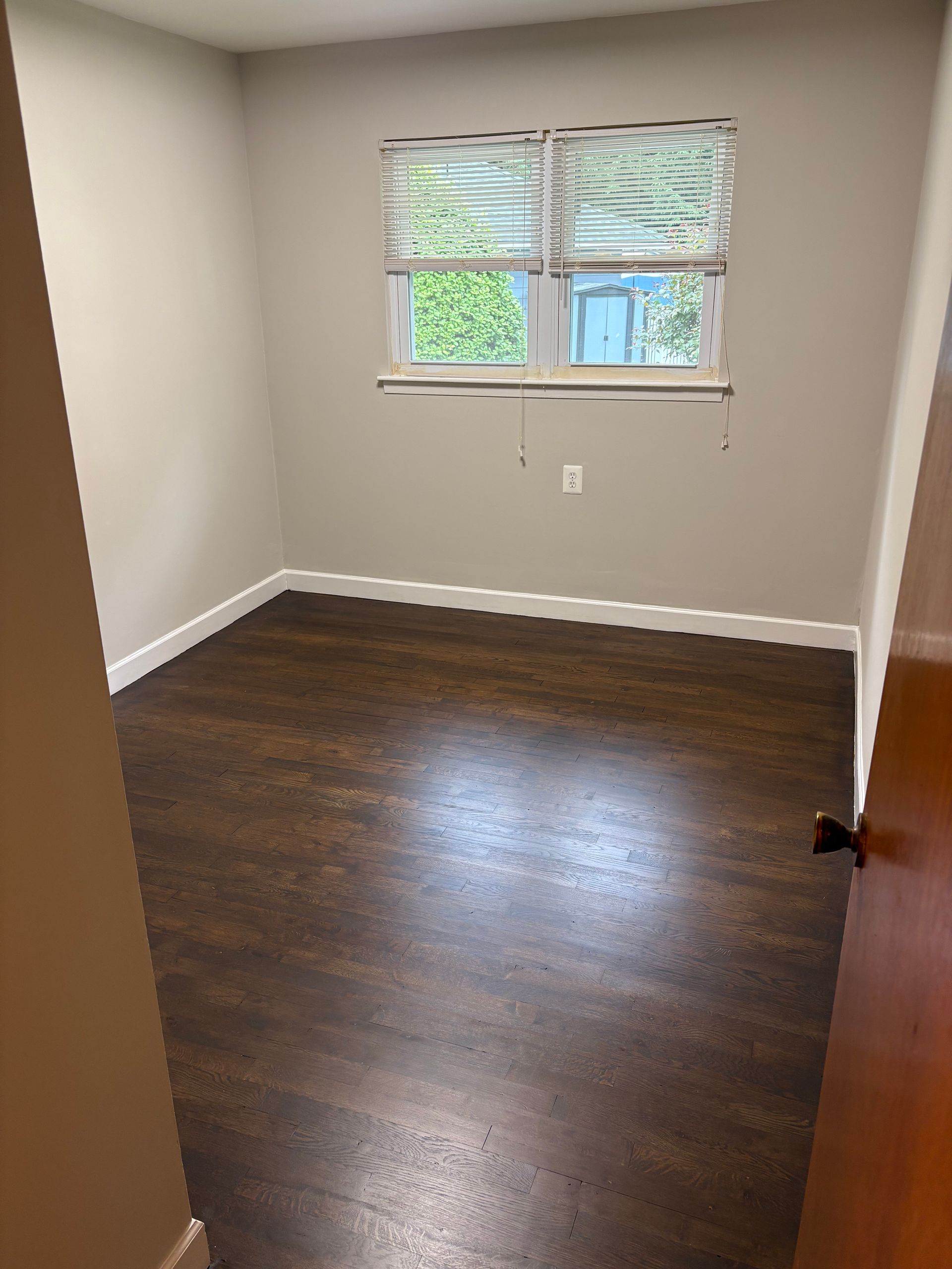 Empty room with dark wood-look flooring, neutral walls, white trim, and a window with blinds.