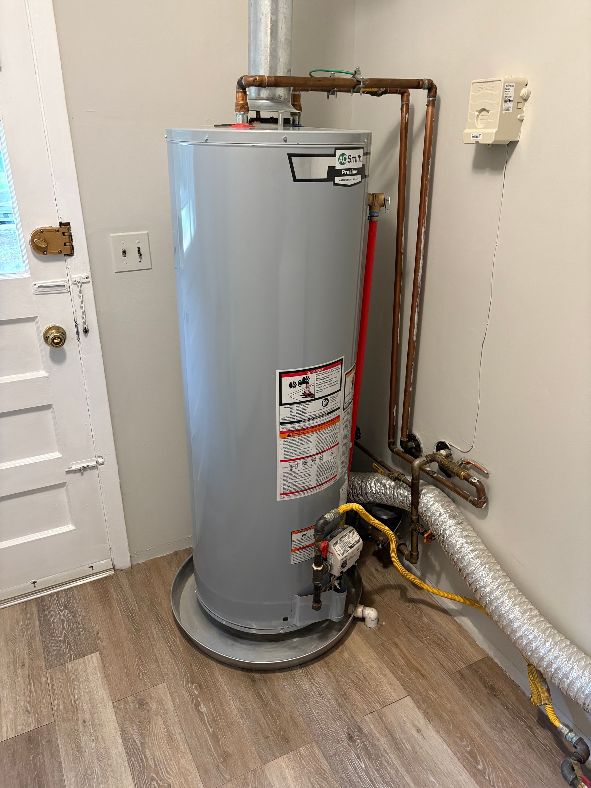 A tall, gray water heater in a room with light-colored flooring and walls. Copper pipes and vents are attached.