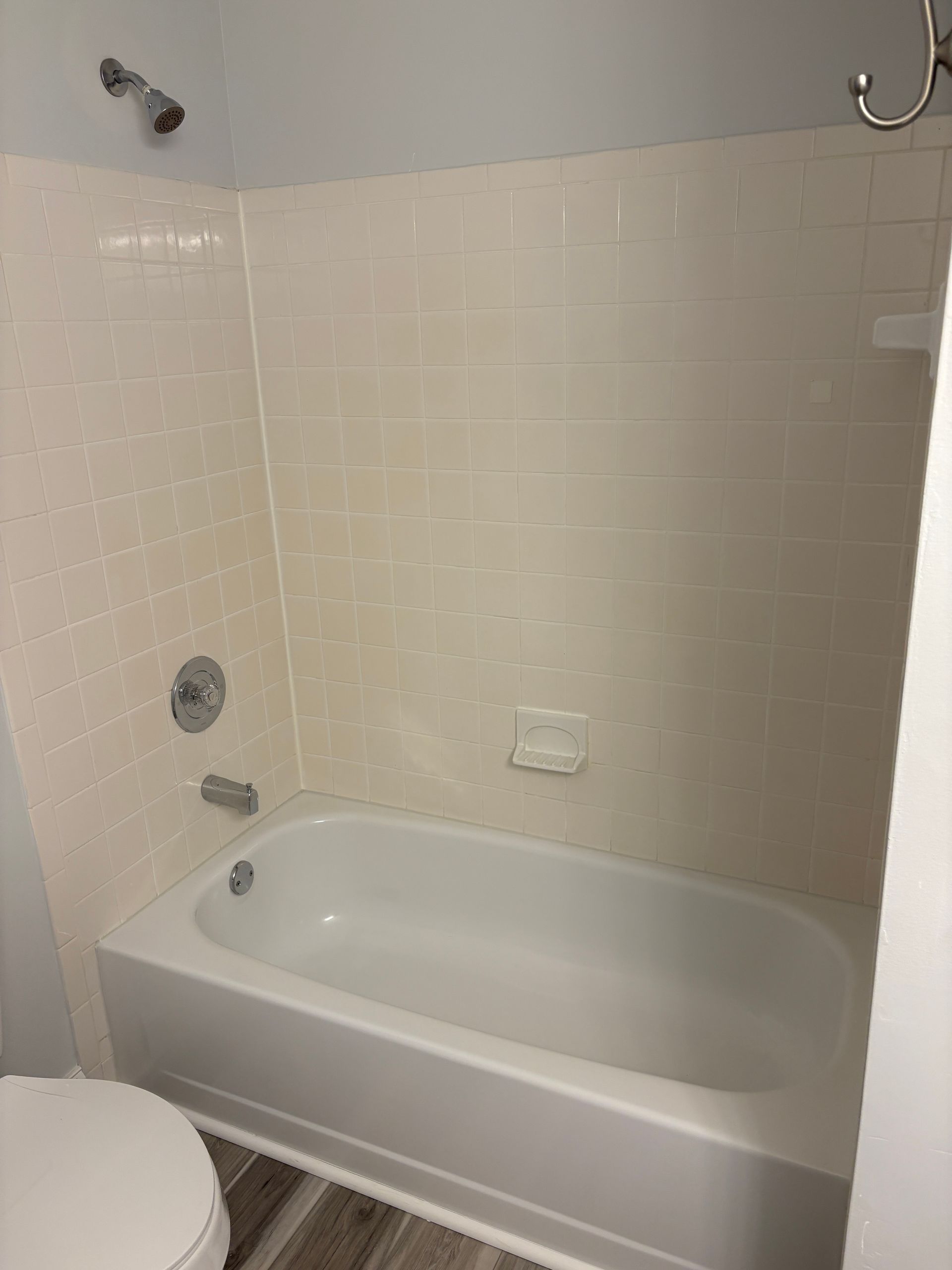 Bathtub with cream-colored tiled walls and fixtures. White tub, chrome faucet, and showerhead. Toilet in foreground.