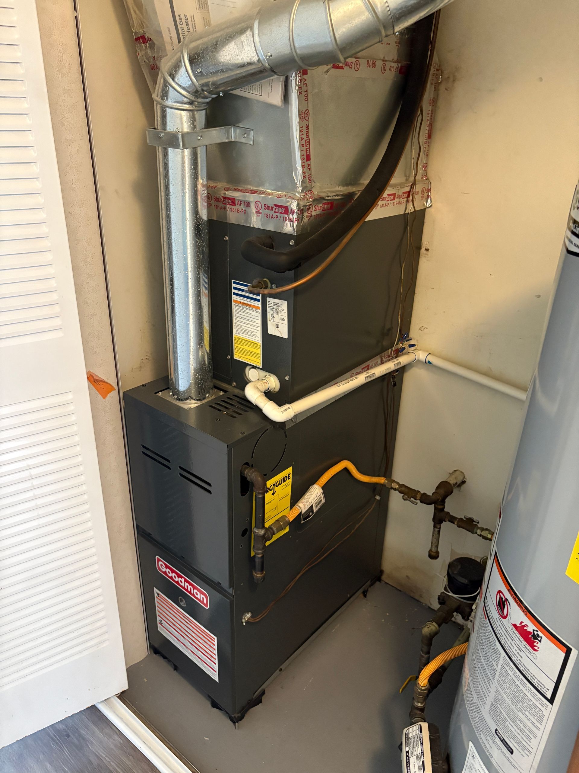 Furnace and water heater in a utility closet. Silver vent, gray units, yellow gas line.
