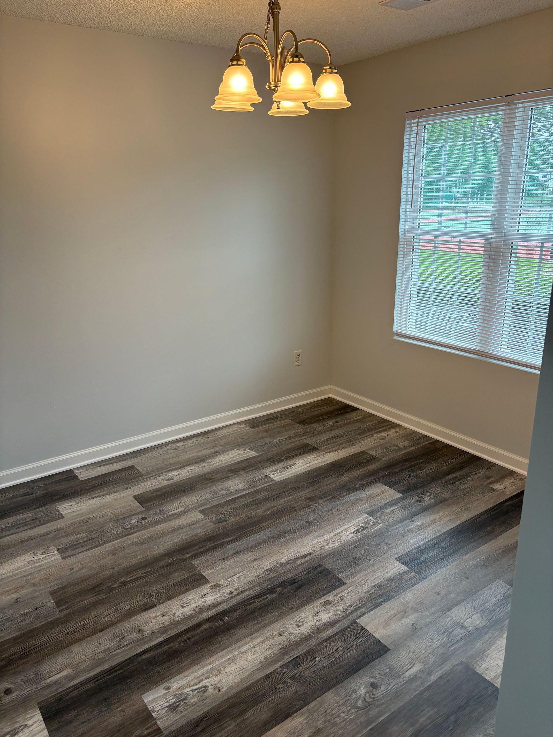 Empty dining room with gray wood-look flooring, beige walls, and a chandelier; a window with blinds is on the right.