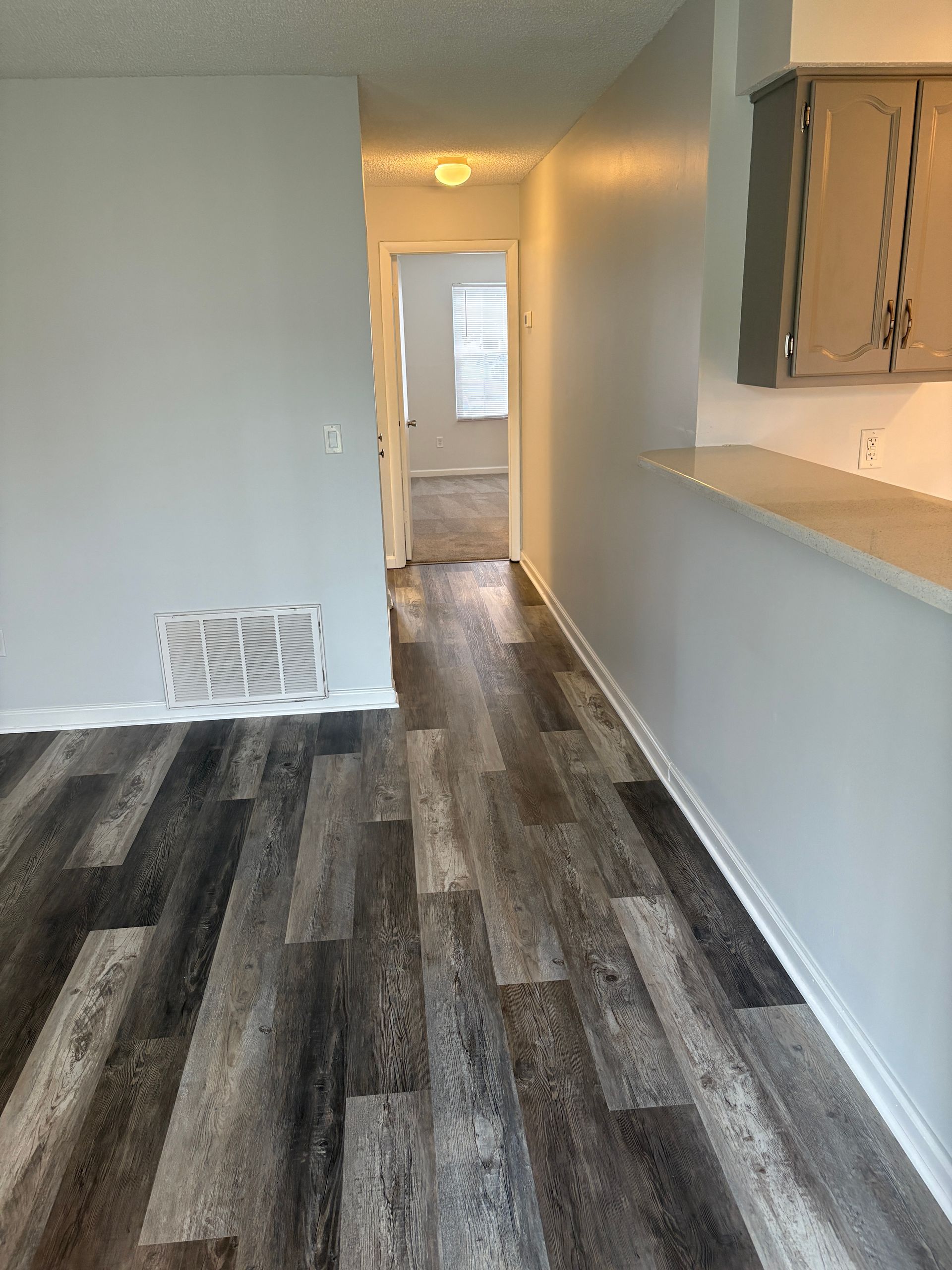 Hallway with dark wood-look flooring, light gray walls, and a view into a room with a window.