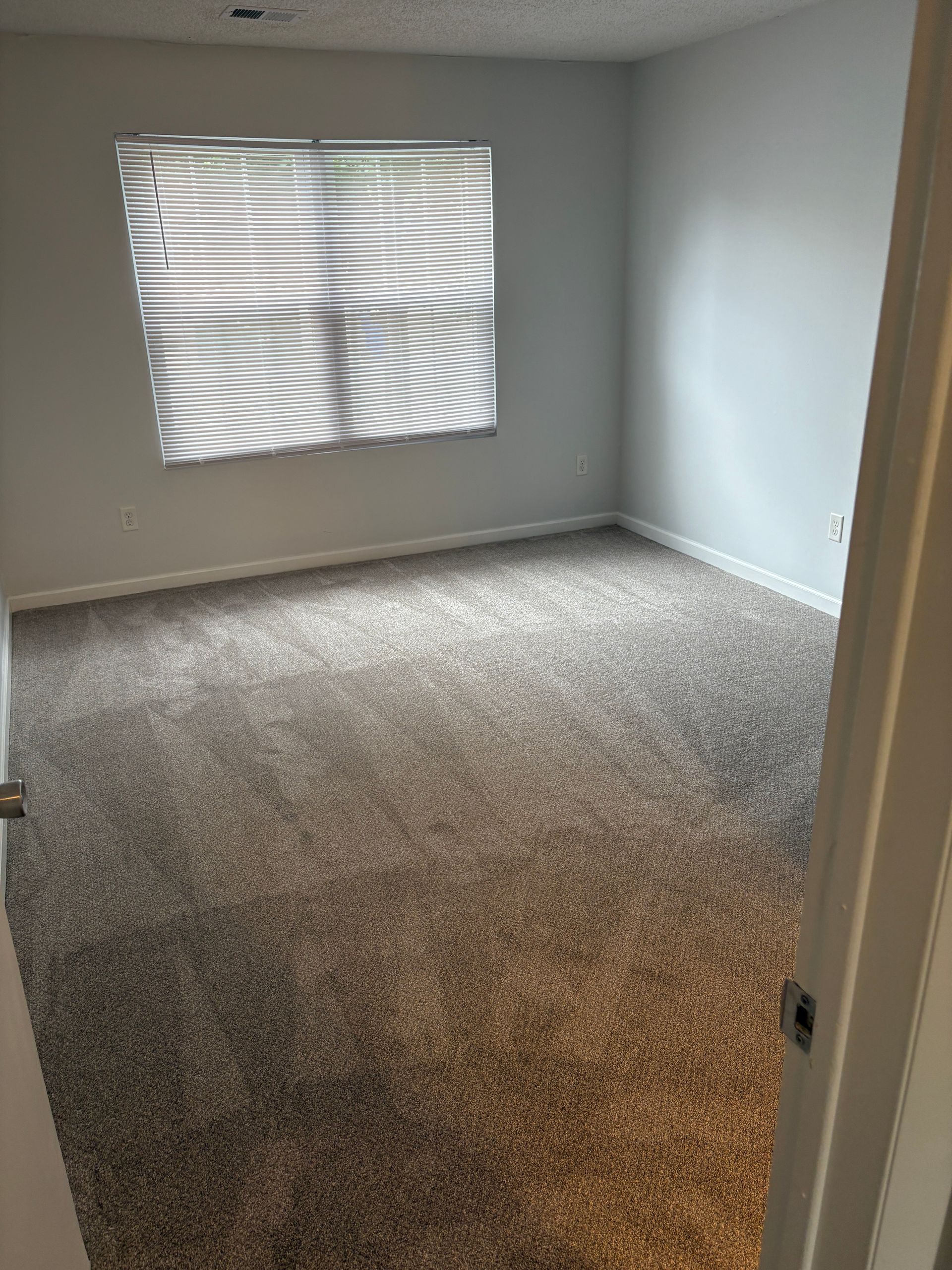 Empty bedroom with gray carpet, blinds on window, light gray walls.