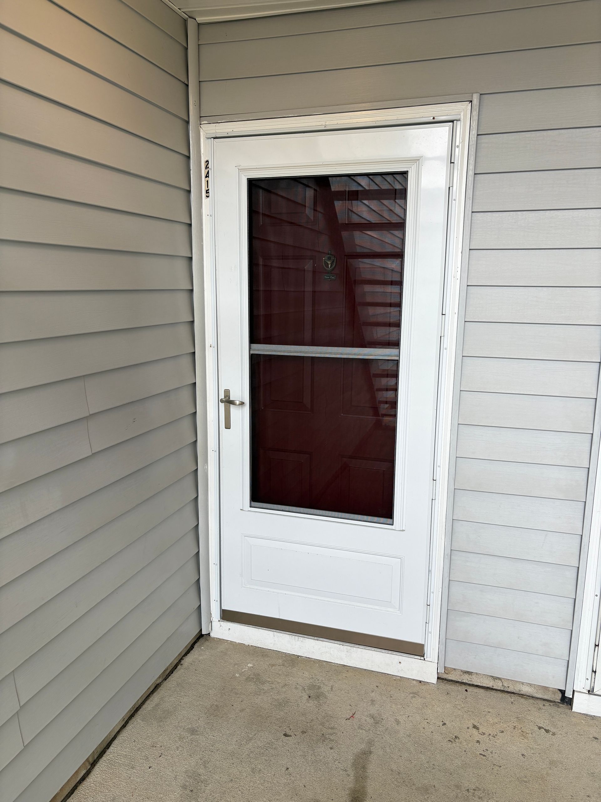 White screen door, gray siding, concrete floor. The door has a silver handle and a dark interior is visible.