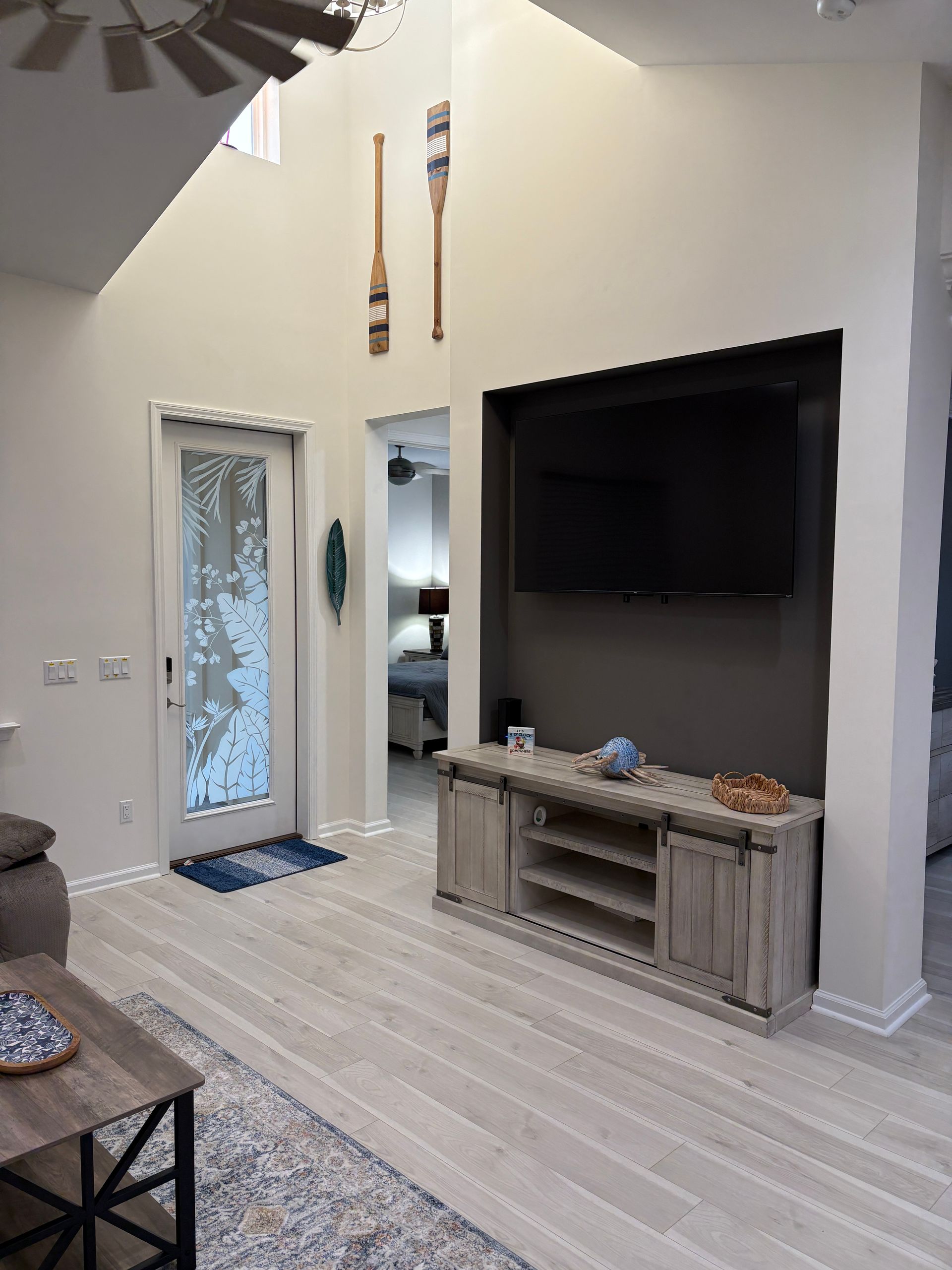 Living room with TV above a wooden cabinet, tall ceiling, oars, and a glass door.