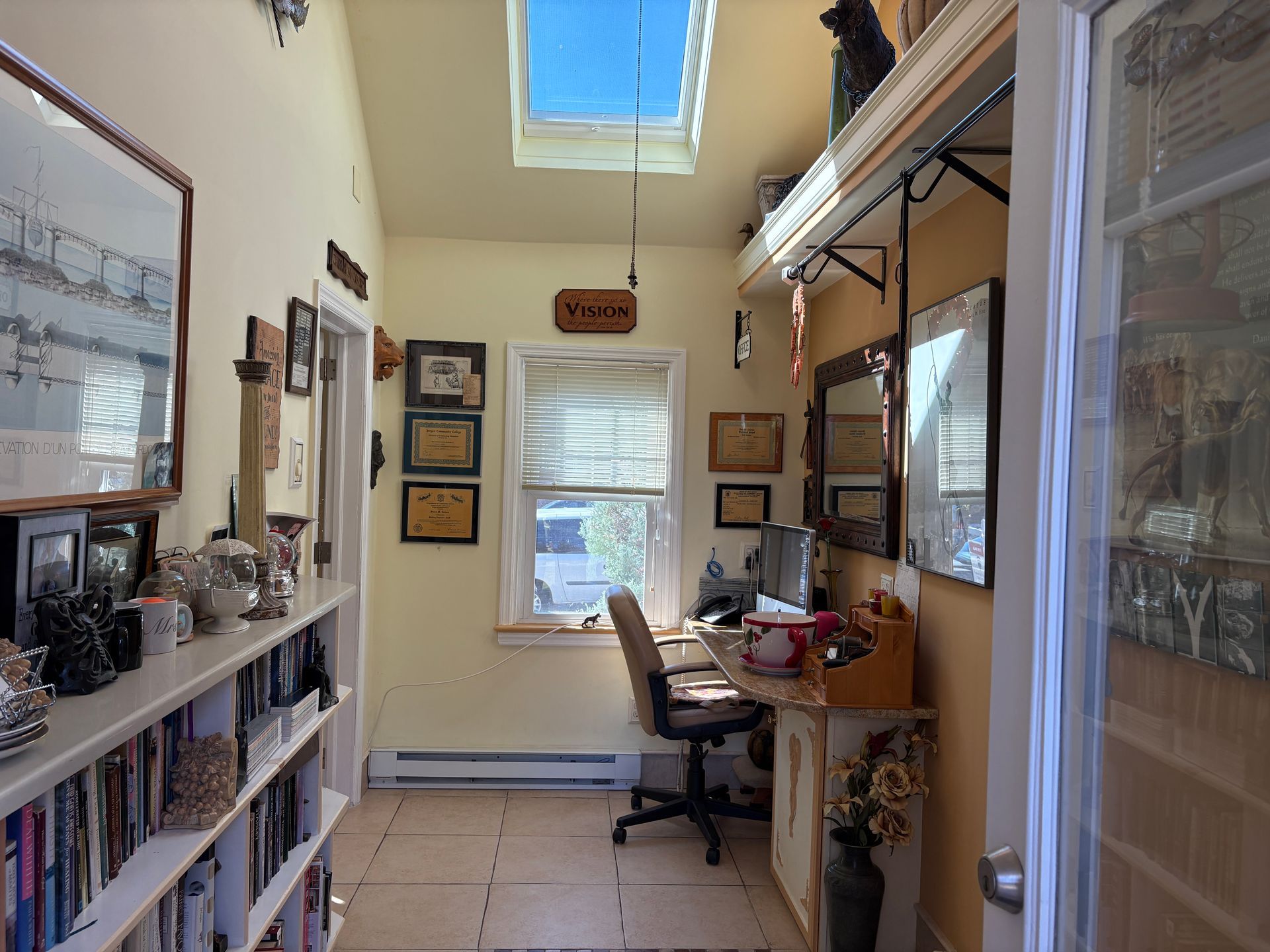 Narrow hallway with home office area. Bookshelves, desk by window under skylight, various decor, and open door.