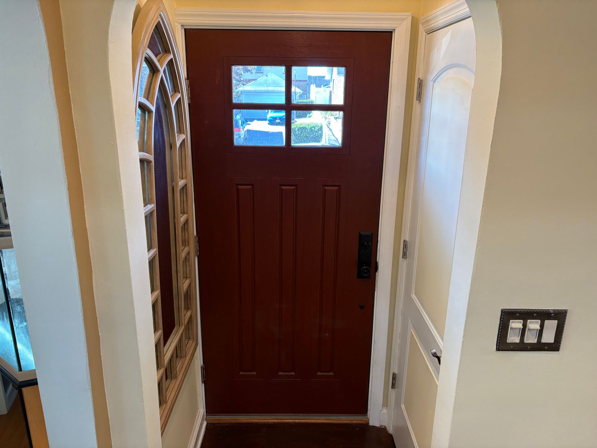 Red front door with glass panes, flanked by an arched window and a wall-mounted light switch.