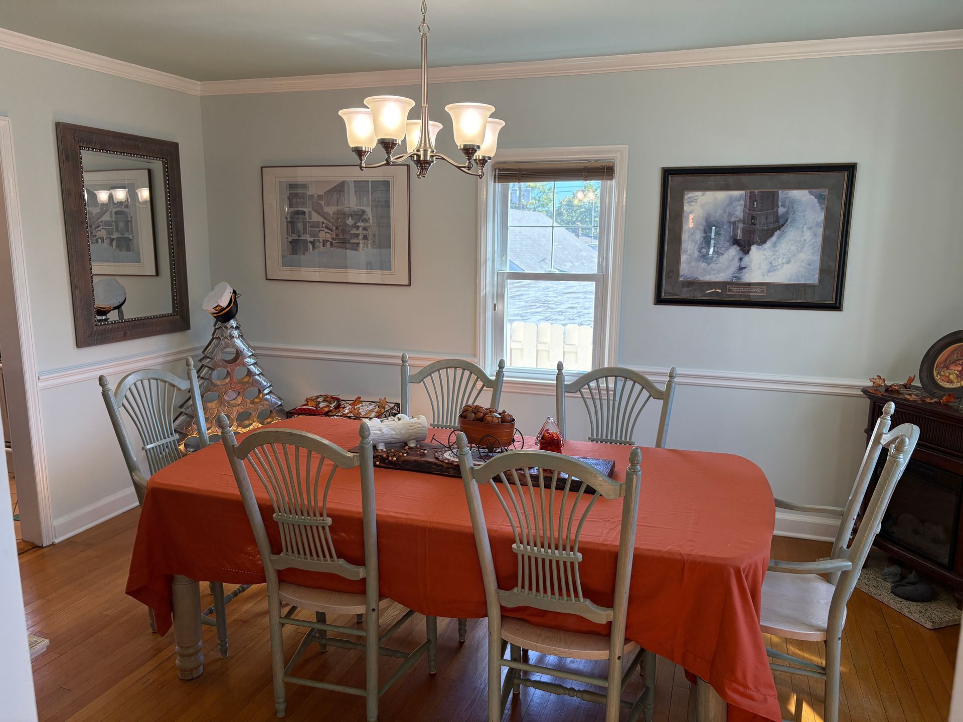 Dining room with an orange tablecloth, wooden chairs, and artwork on light blue walls.