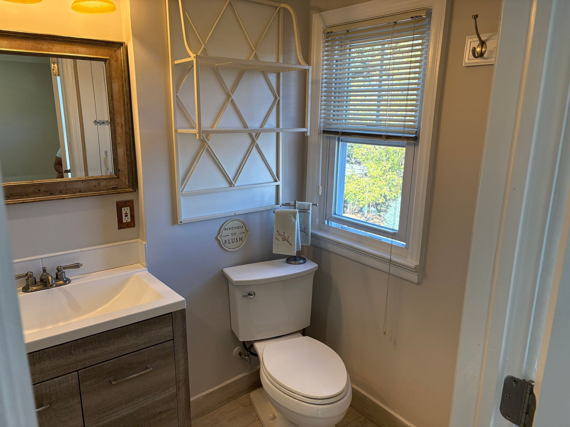 Small bathroom with a toilet, sink, mirror, window, and decorative shelf.