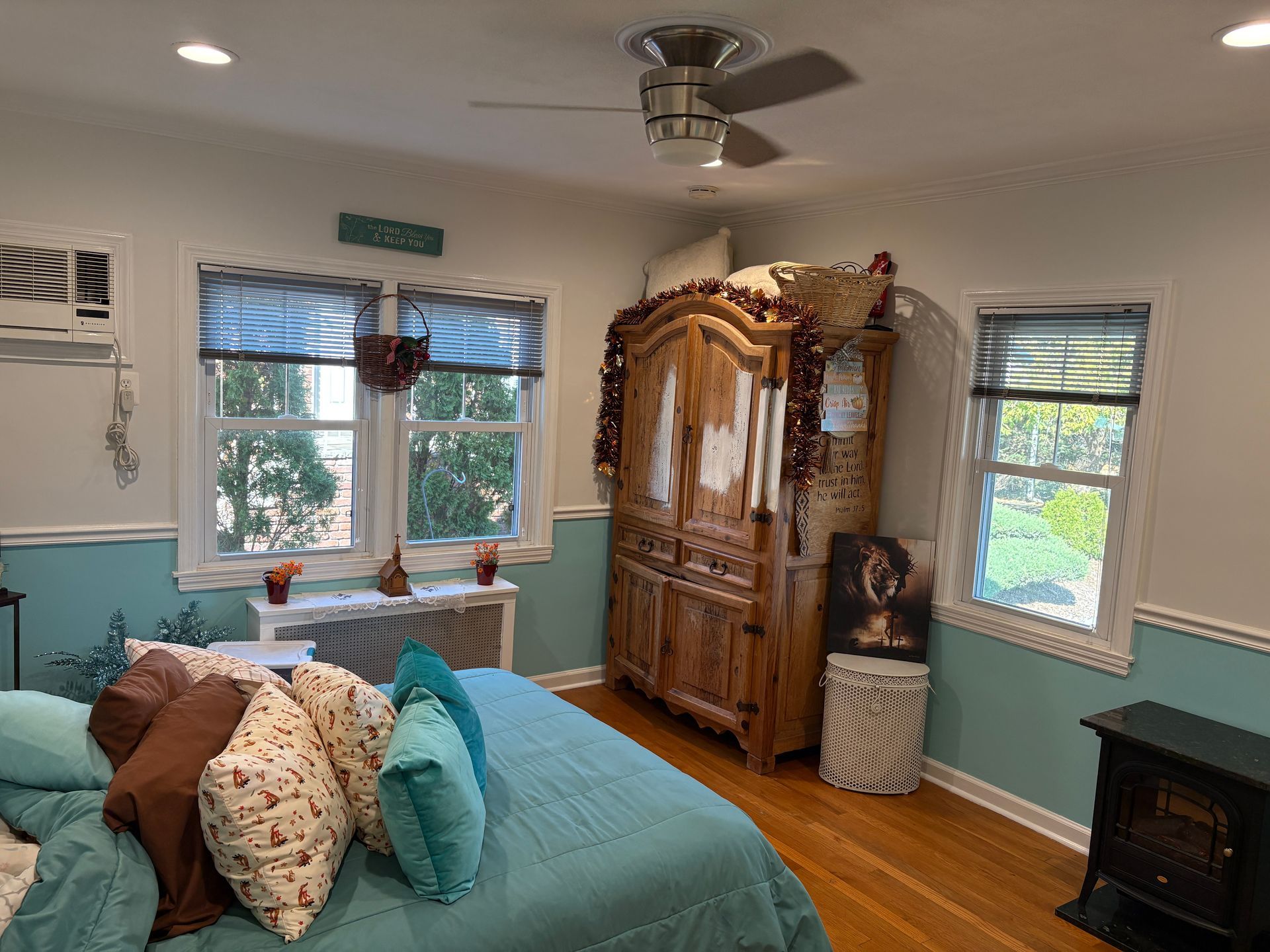 Bedroom with a large wooden cabinet, bed with turquoise bedding and pillows, two windows.