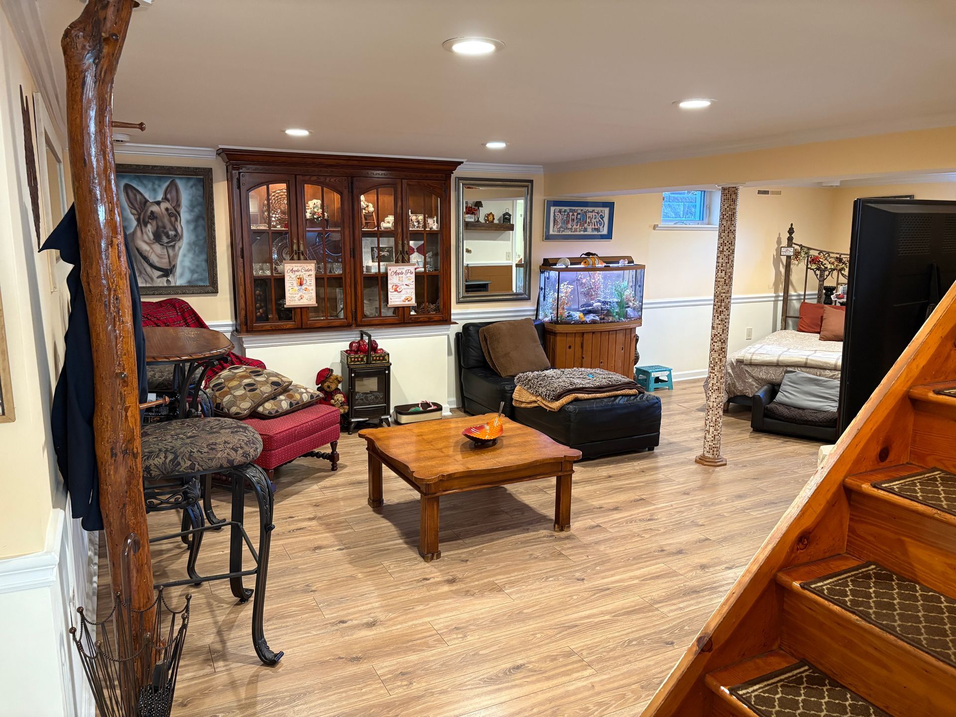 Basement living area: wood floor, stairs, furniture, large cabinet, aquarium, dog portrait on wall.