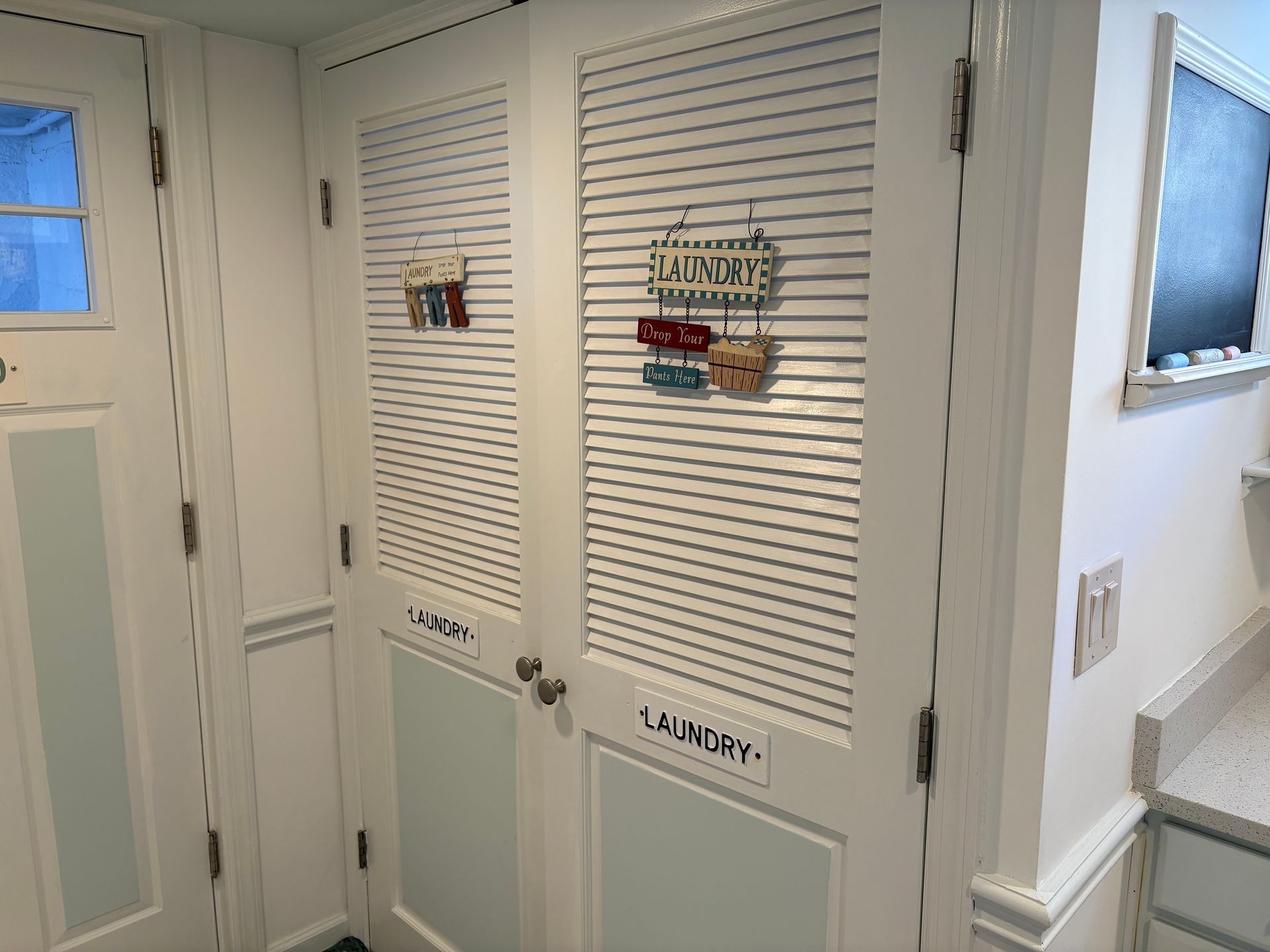 Laundry room with white louvered doors, decorative signs, and a window.