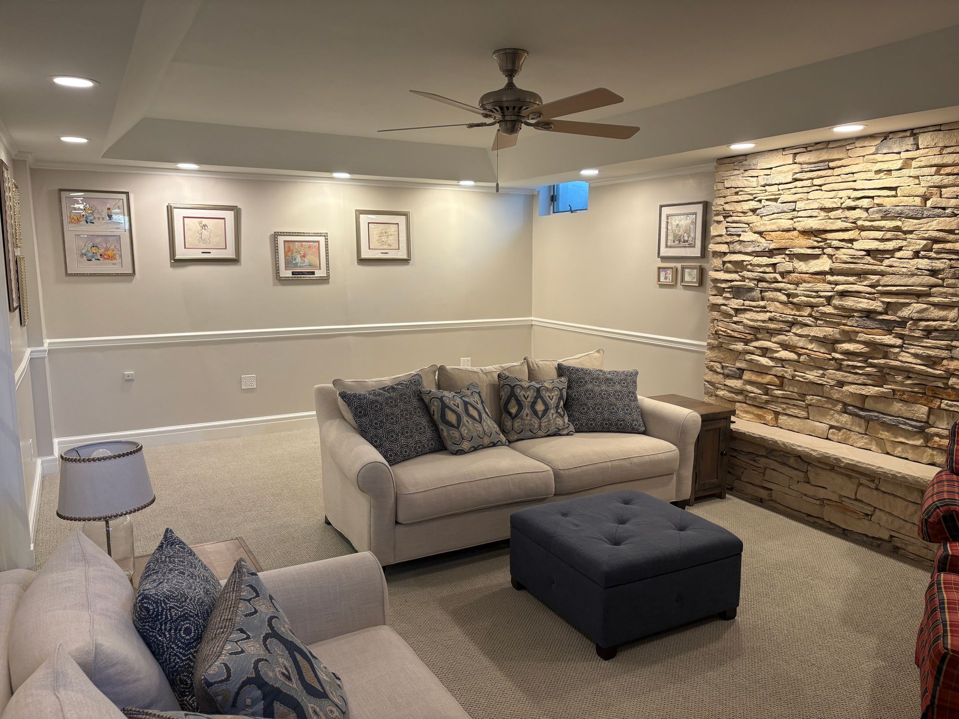 Cozy living room with cream sofa, blue pillows, patterned carpet, stone wall, and framed artwork.