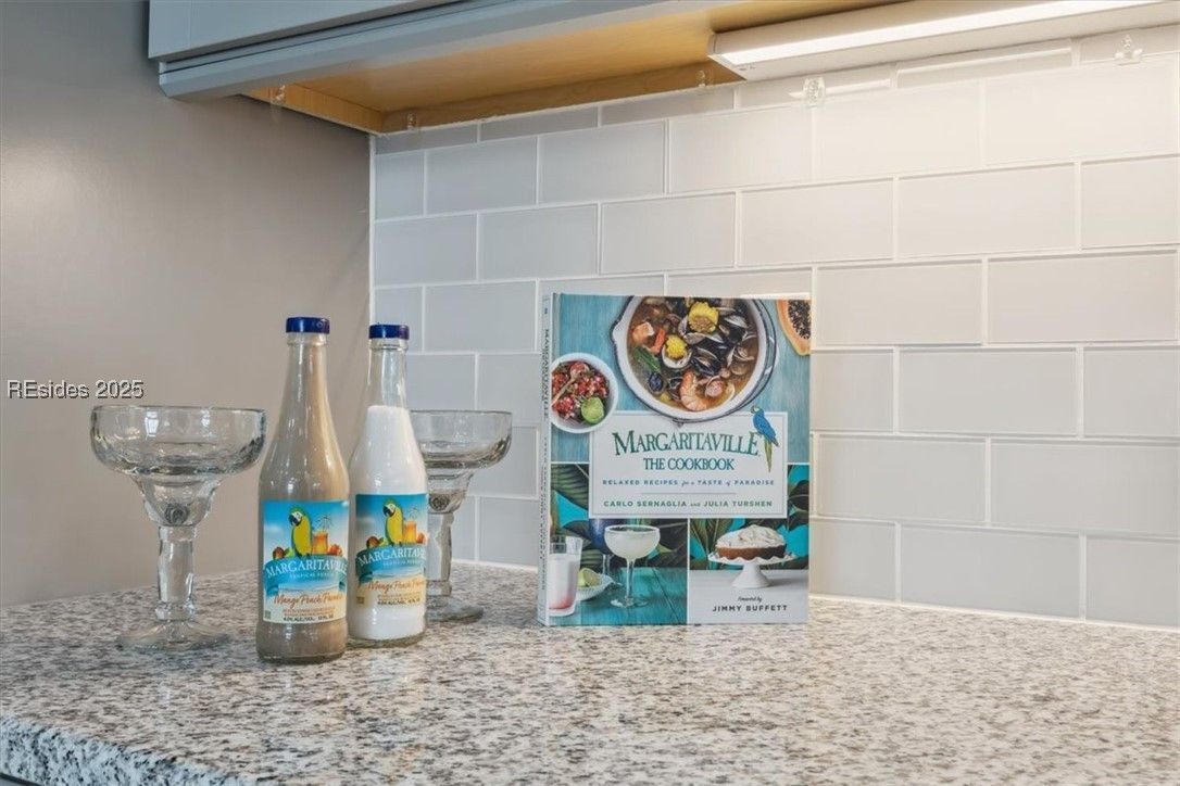 Bar setup: margarita glasses, bottles, cookbook on a speckled countertop against a tiled backsplash.