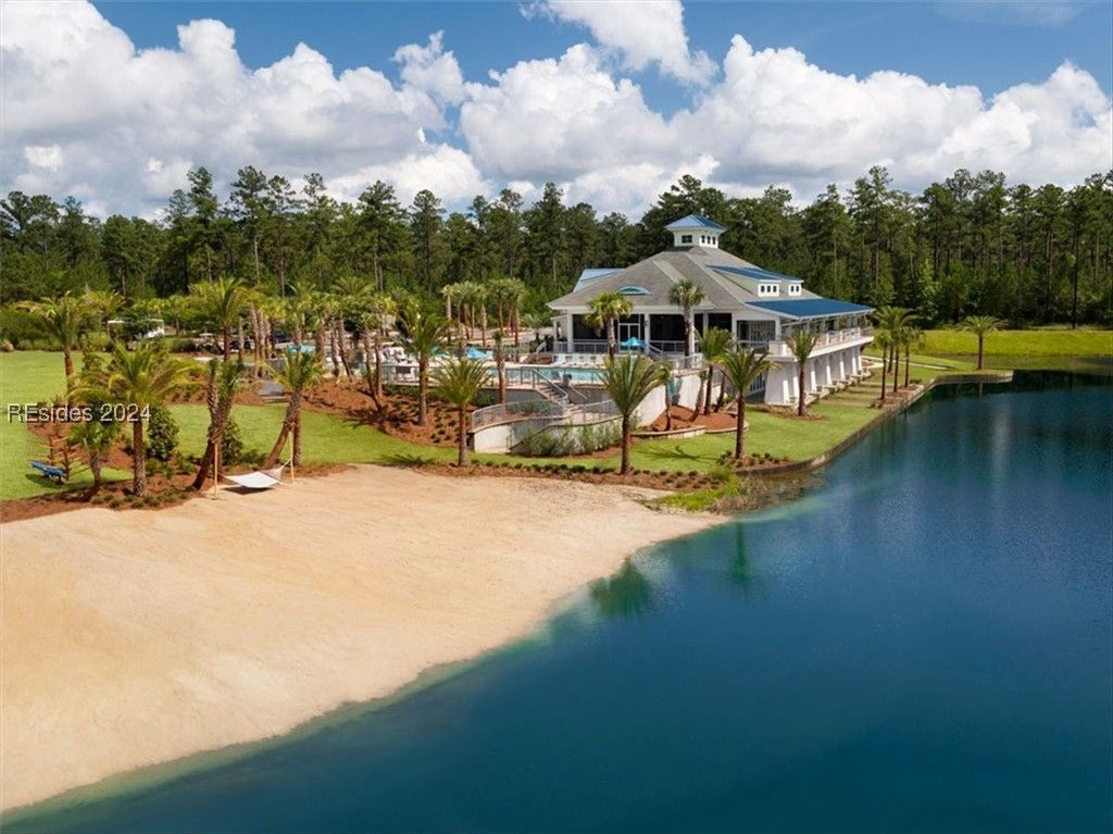 Beachfront clubhouse with a pool and sandy shore, set against a lake and trees, under a blue sky.