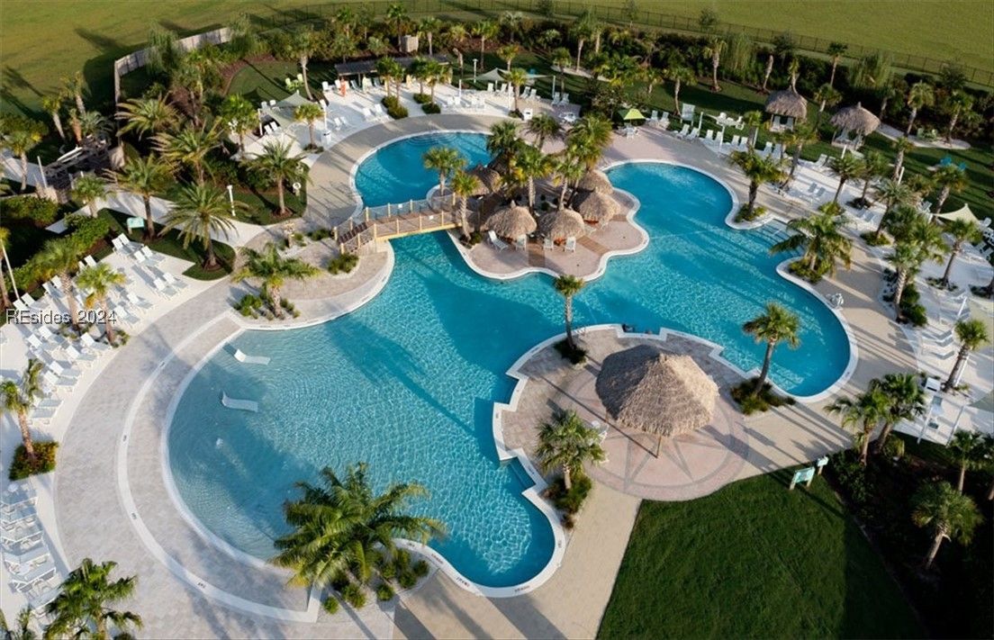 Aerial view of a large, multi-shaped blue pool with bridges and tiki huts, surrounded by palm trees and a white deck.