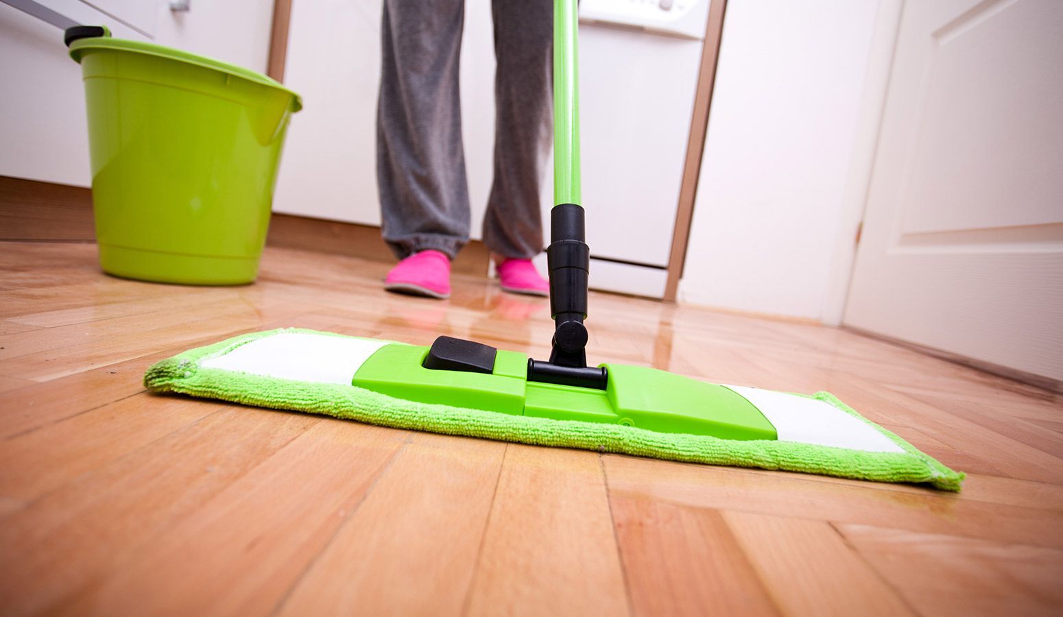 Person mopping a wooden floor with a green mop, green bucket, and wearing gray pants and pink shoes.