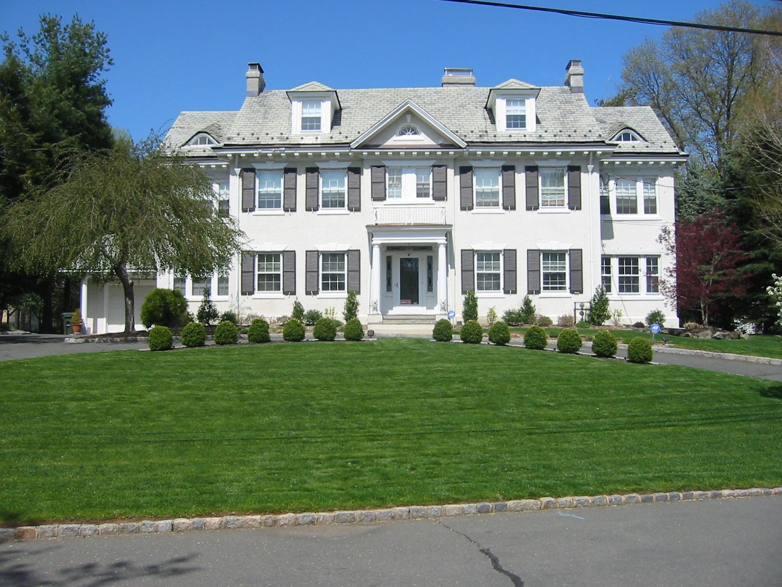 Large white house with gray roof, black shutters, and well-manicured lawn on a sunny day.